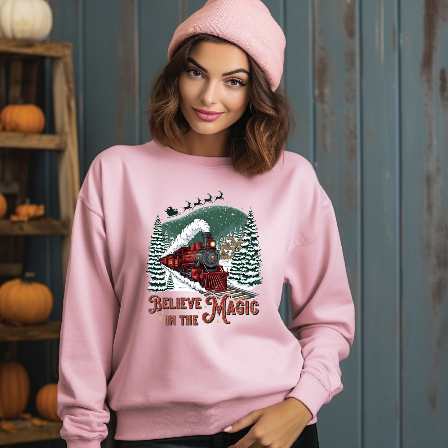 Woman wearing a pink sweatshirt with a Christmas design and text, standing against a blue wall with pumpkins on shelves.