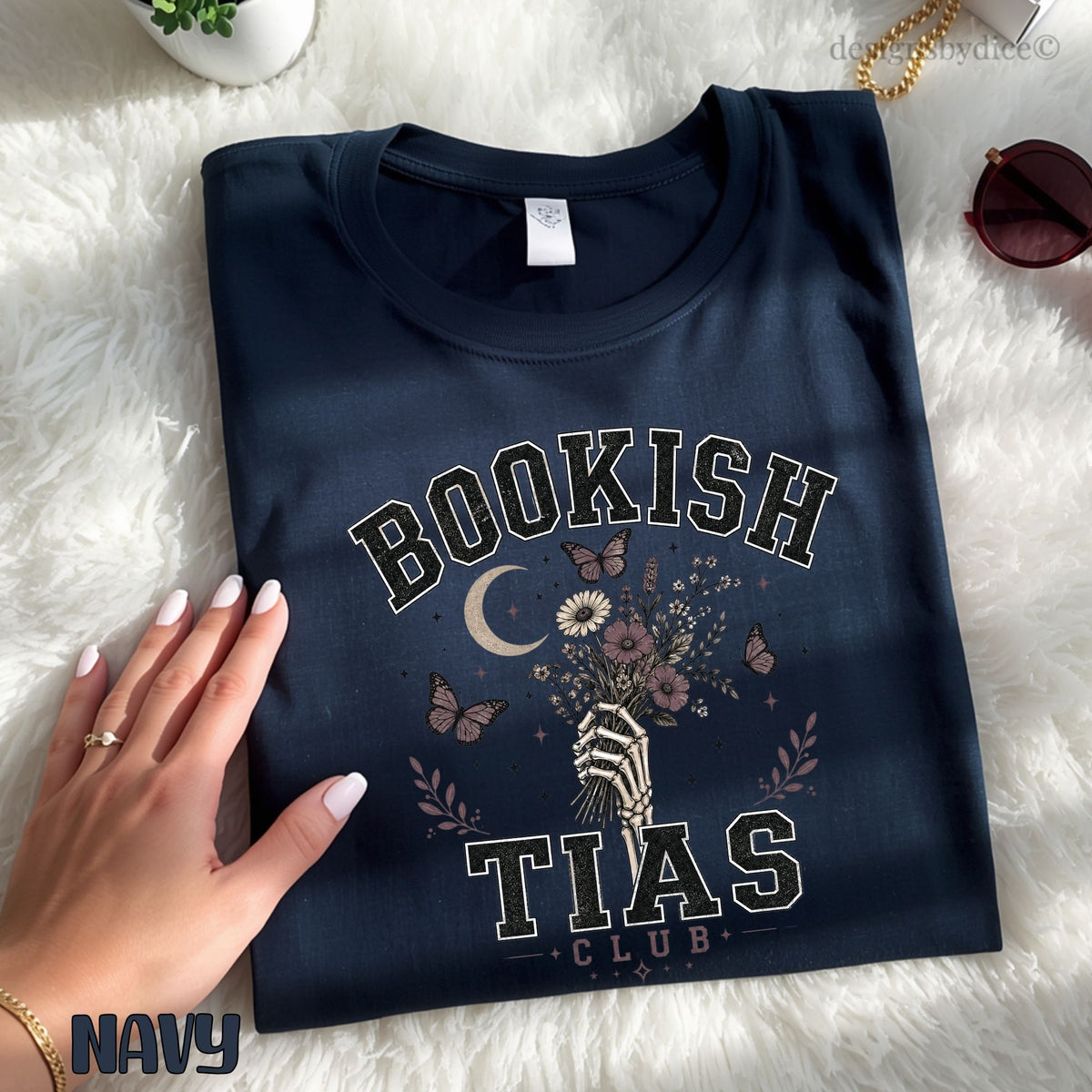 A navy blue folded graphic T-shirt laid flat on a white faux-fur surface, with a hand resting on the left side. The shirt features a vintage-style print reading “BOOKISH TIAS CLUB” with a hand holding flowers, butterflies, a crescent moon, and floral details.