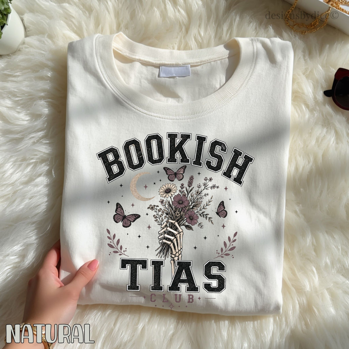 A folded cream-colored sweatshirt with a graphic print reading 'BOOKISH TIAS CLUB' and featuring a hand holding a bouquet of flowers with butterflies, laid flat on a white faux fur surface. A hand is holding the left side of the sweatshirt.