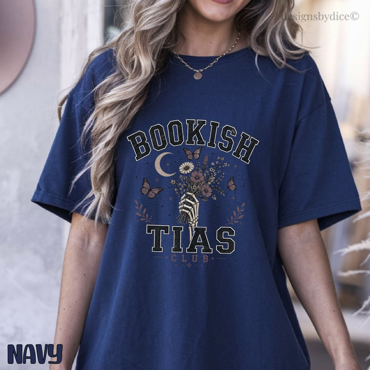 A woman wearing an oversized navy blue graphic T-shirt with the words 'BOOKISH TIAS CLUB' and a floral/butterfly print on the front. The photo is cropped from the lower face to the hips, with a softly blurred indoor background.