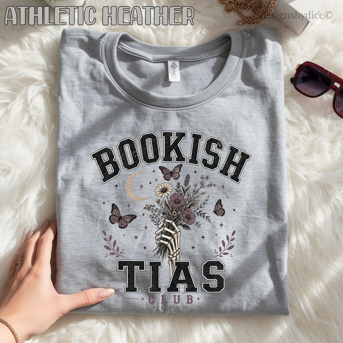 A folded heather gray graphic T-shirt laid flat on a white faux-fur surface, with a hand holding one corner. The shirt has a large vintage-style black and muted purple graphic reading “BOOKISH TIAS CLUB” with butterflies, flowers, and a striped hand holding a bouquet.