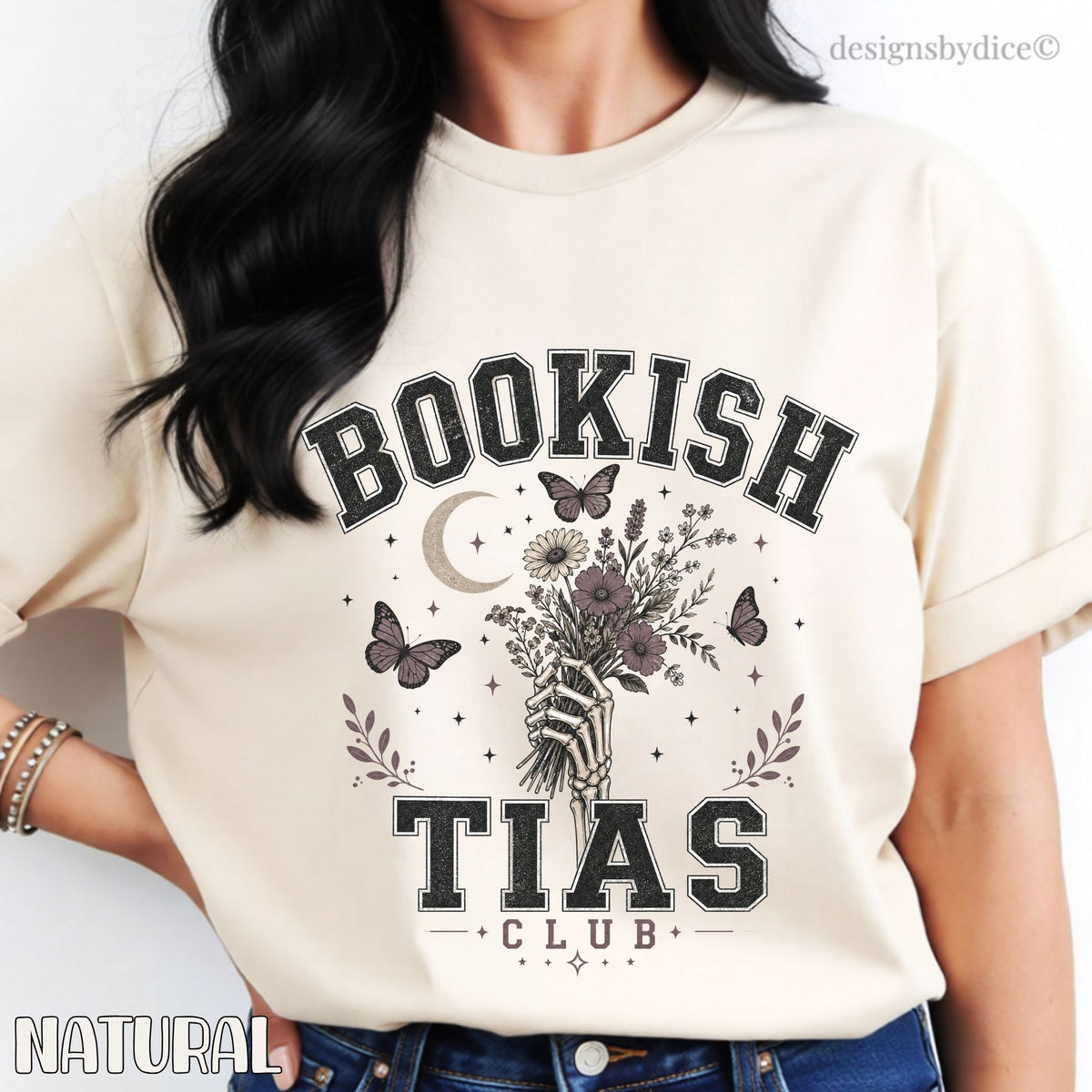 A woman wearing a cream-colored graphic t-shirt with rolled sleeves. The shirt features a gothic-style print that reads 'BOOKISH TIAS CLUB' with an illustration of a skeleton hand holding a bouquet of flowers, surrounded by butterflies, a crescent moon, and stars.