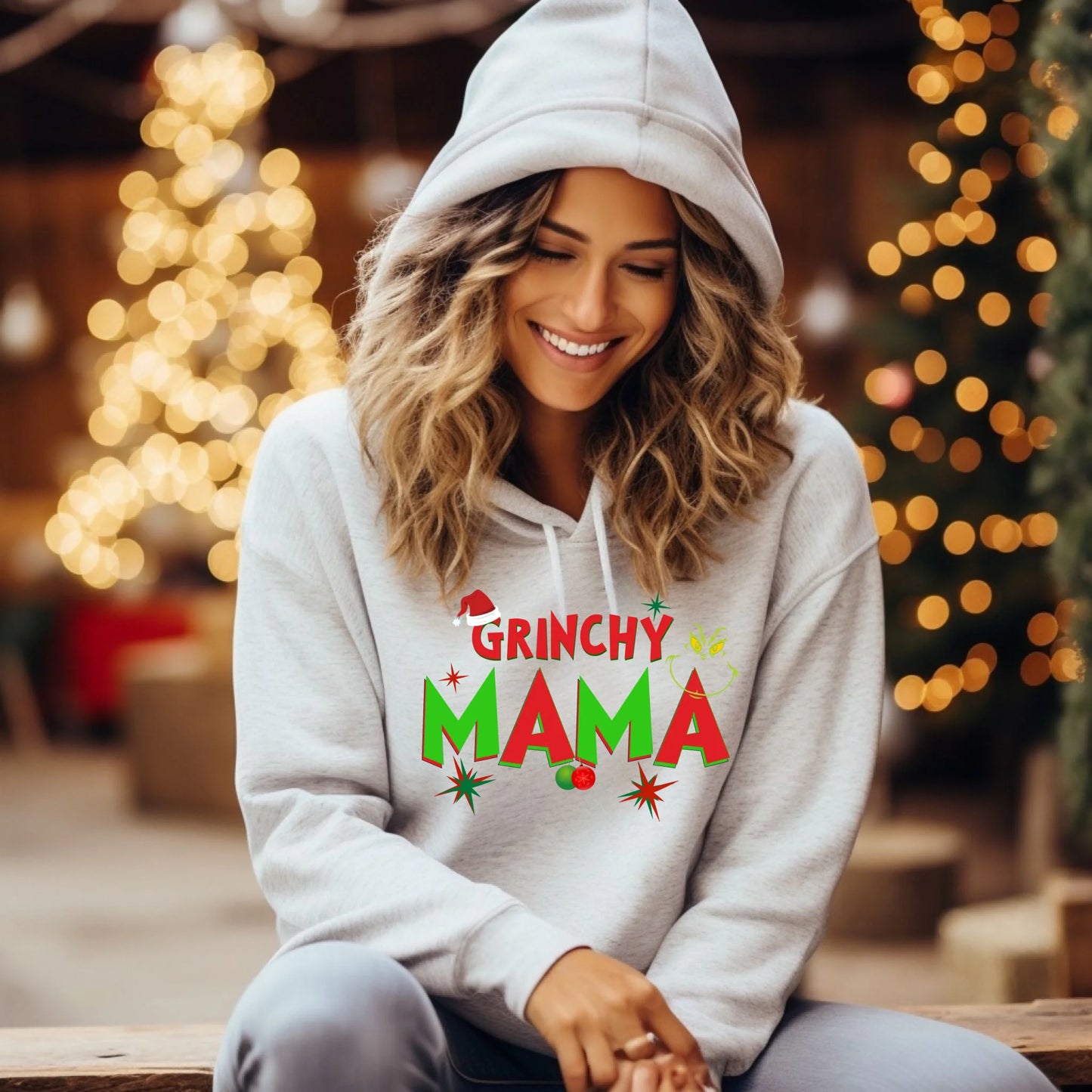 Woman wearing a 'Grinchy Mama' hoodie with festive design, sitting in a decorated indoor setting.
