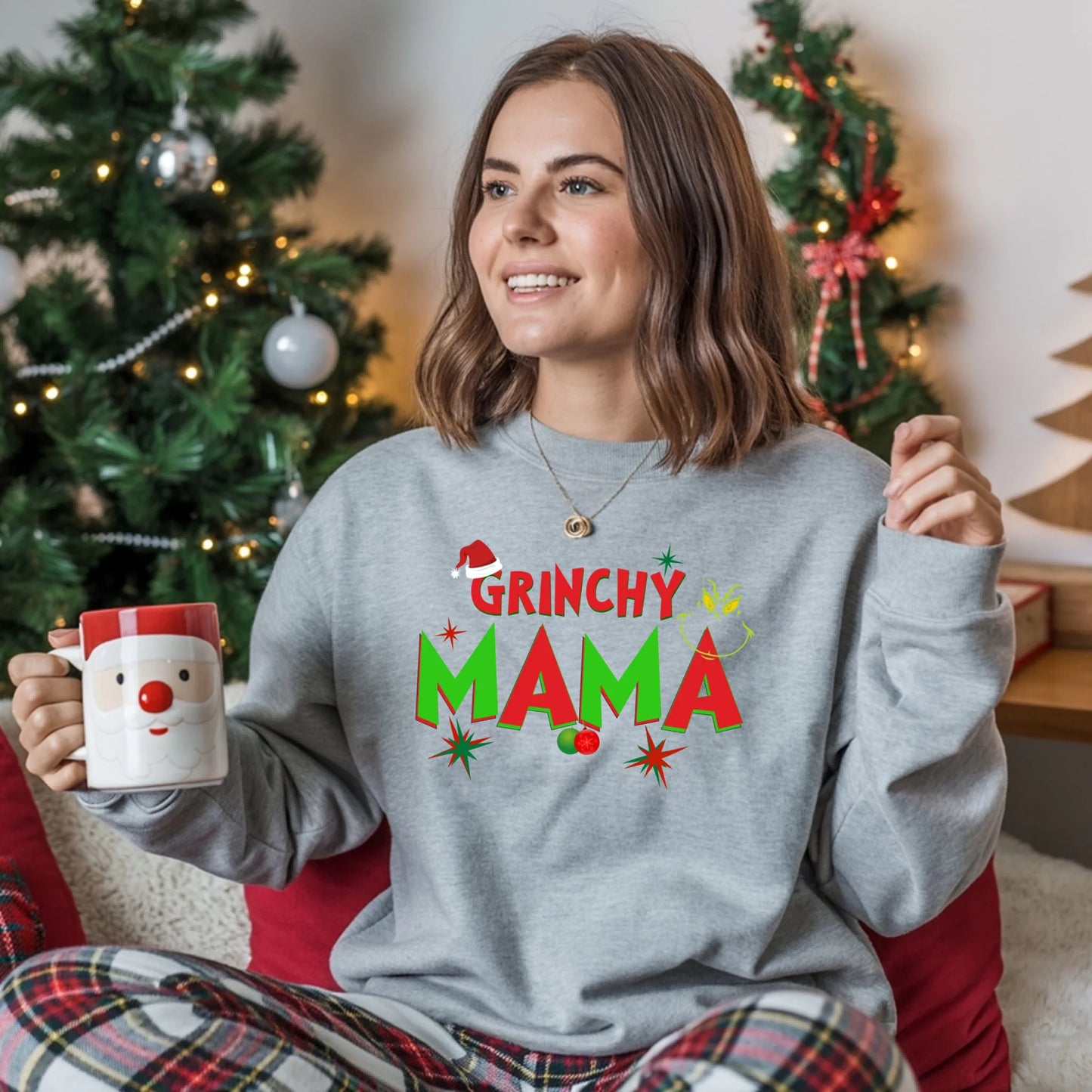 Woman wearing a 'Grinchy Mama' sweater holding a mug in front of a decorated Christmas tree.