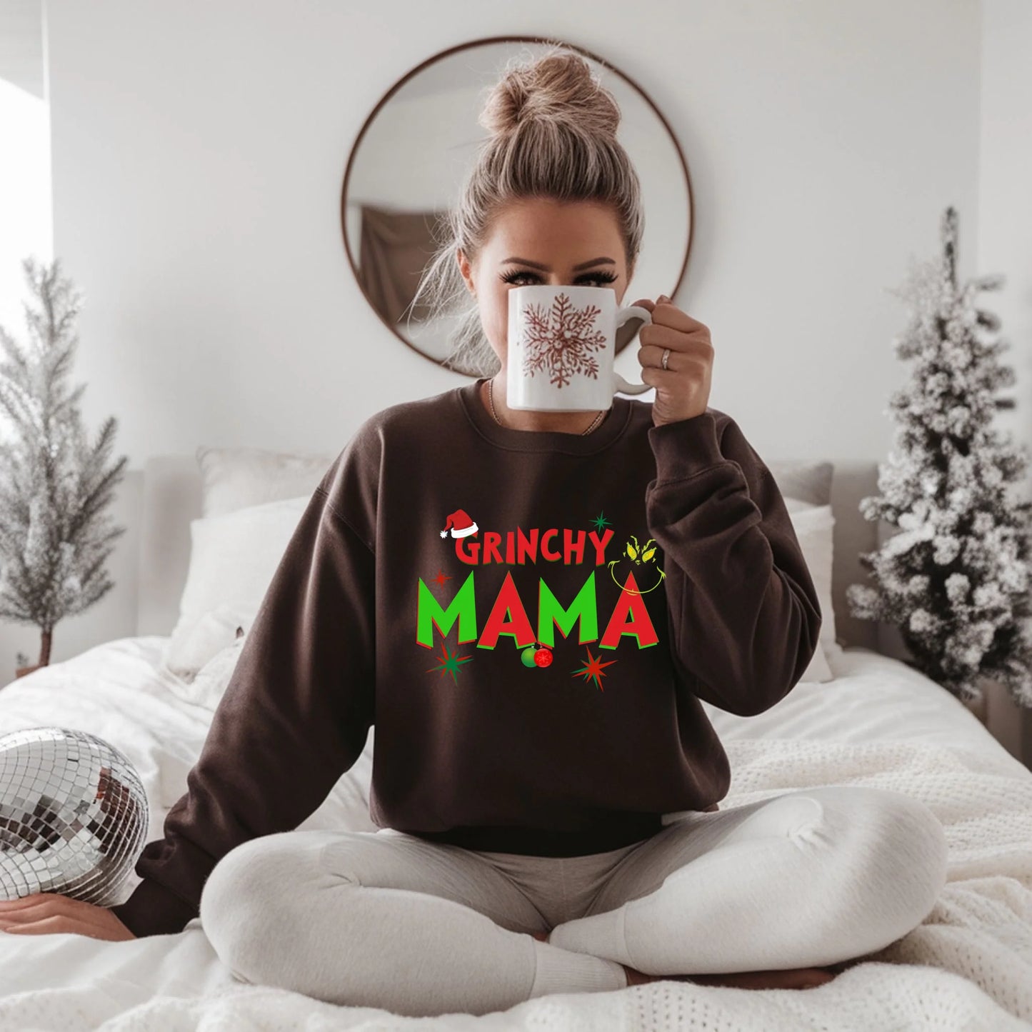 Woman wearing a 'Grinchy Mama' sweater holding a mug in a cozy room.