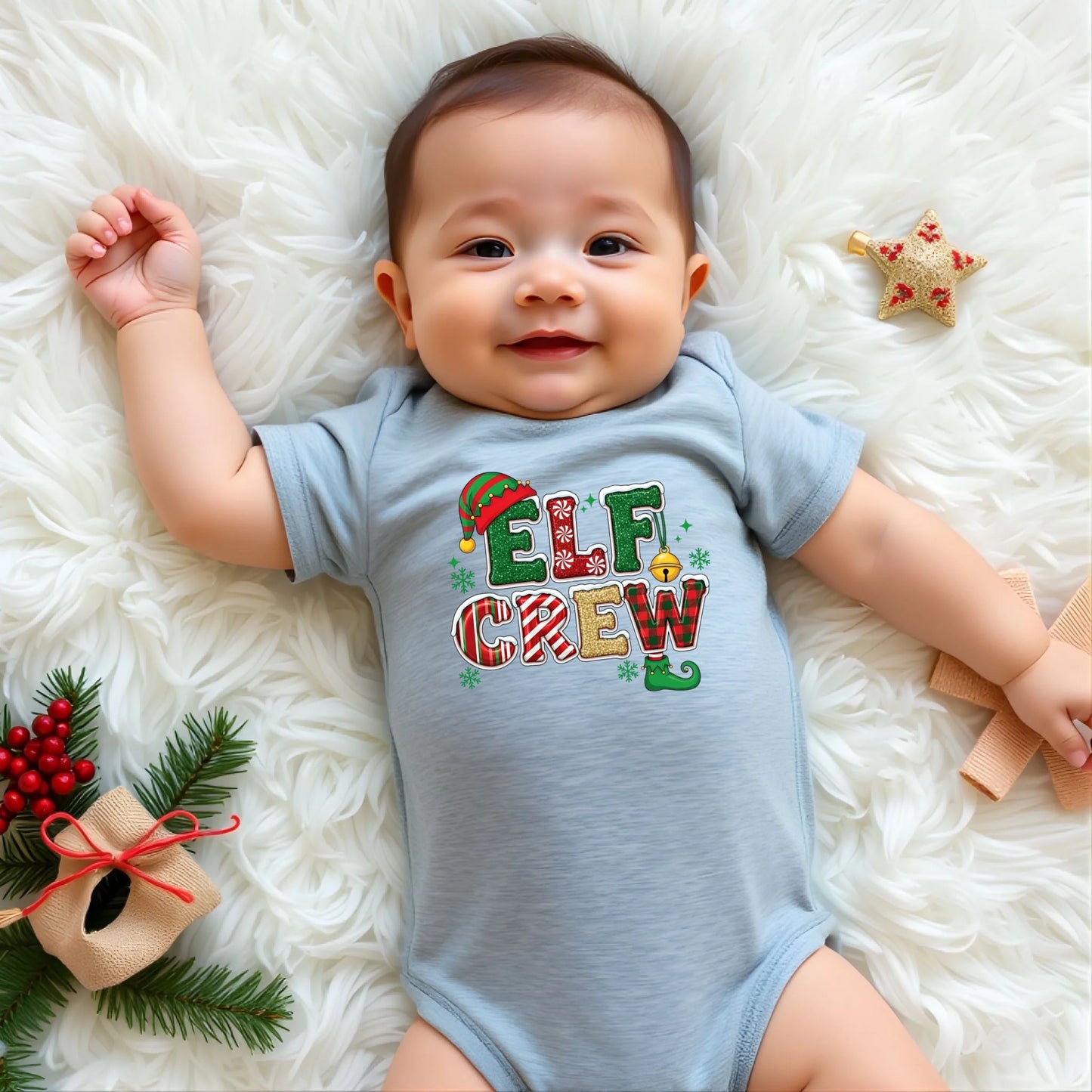 Baby wearing a blue onesie with 'Elf Crew' text, lying on a white fluffy surface with Christmas decorations.