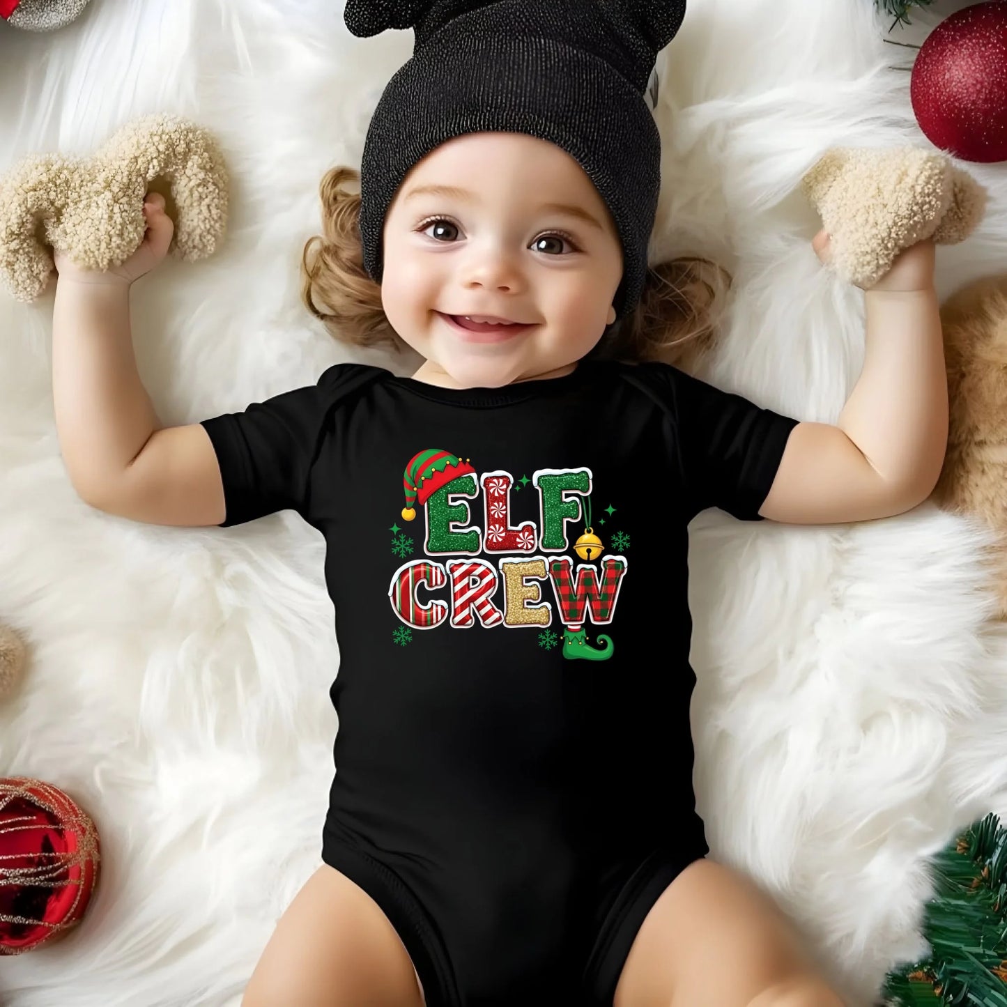 Child wearing a black 'Elf Crew' onesie on a white fluffy surface with Christmas decorations.