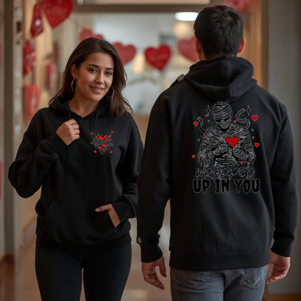Man and woman wearing black hoodies with front heart print and large mummy artwork on the back that reads Wrapped Up In You, romantic Valentine hoodie.
