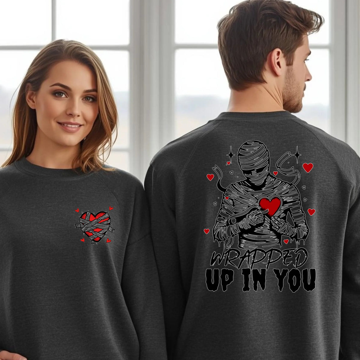 A couple wearing matching charcoal grey sweaters with the Wrapped Up In You mummy and heart design printed on the back, romantic gift apparel.