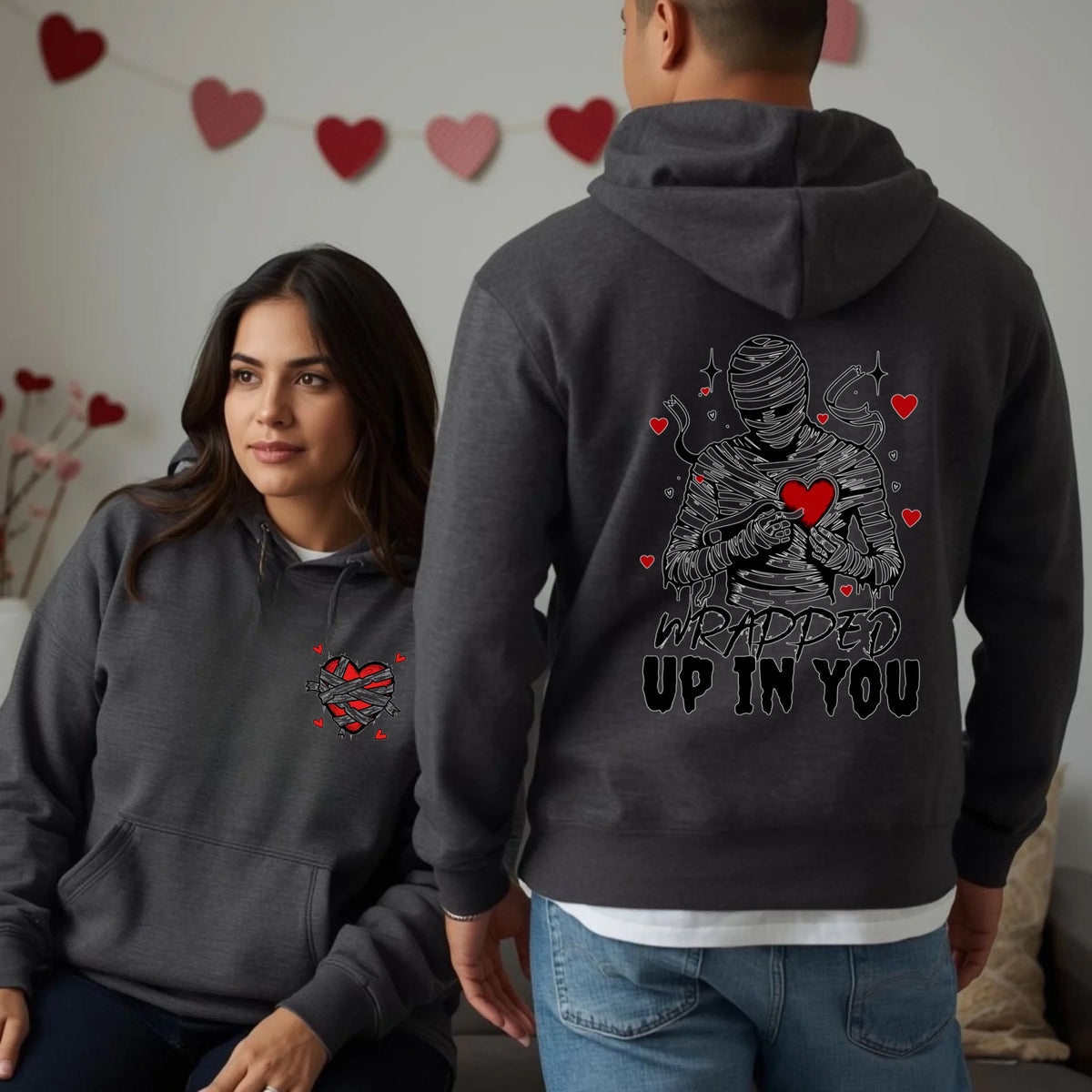 Matching charcoal grey hoodies with heart front detail and Wrapped Up In You mummy design on the back, worn by a couple in a Valentine setting.