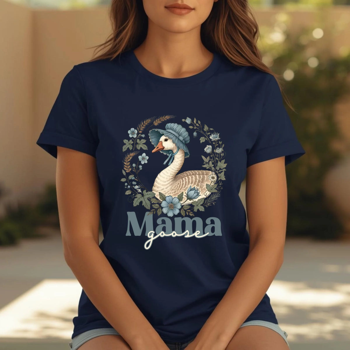 Woman seated outdoors wearing a navy Mama Goose motherhood t-shirt featuring a hand-drawn goose and floral wreath design, relaxed and natural pose.
