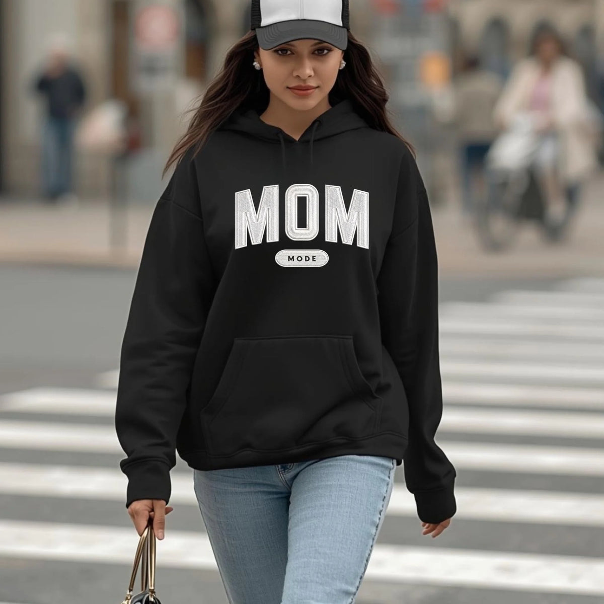 Woman wearing a black hoodie with 'MOM' embroidered printed on it, walking on a crosswalk.