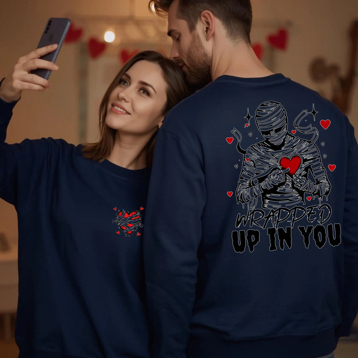 Woman and man taking a selfie wearing navy crewneck sweatshirts featuring a mummy hugging a heart and Wrapped Up In You text on the back.