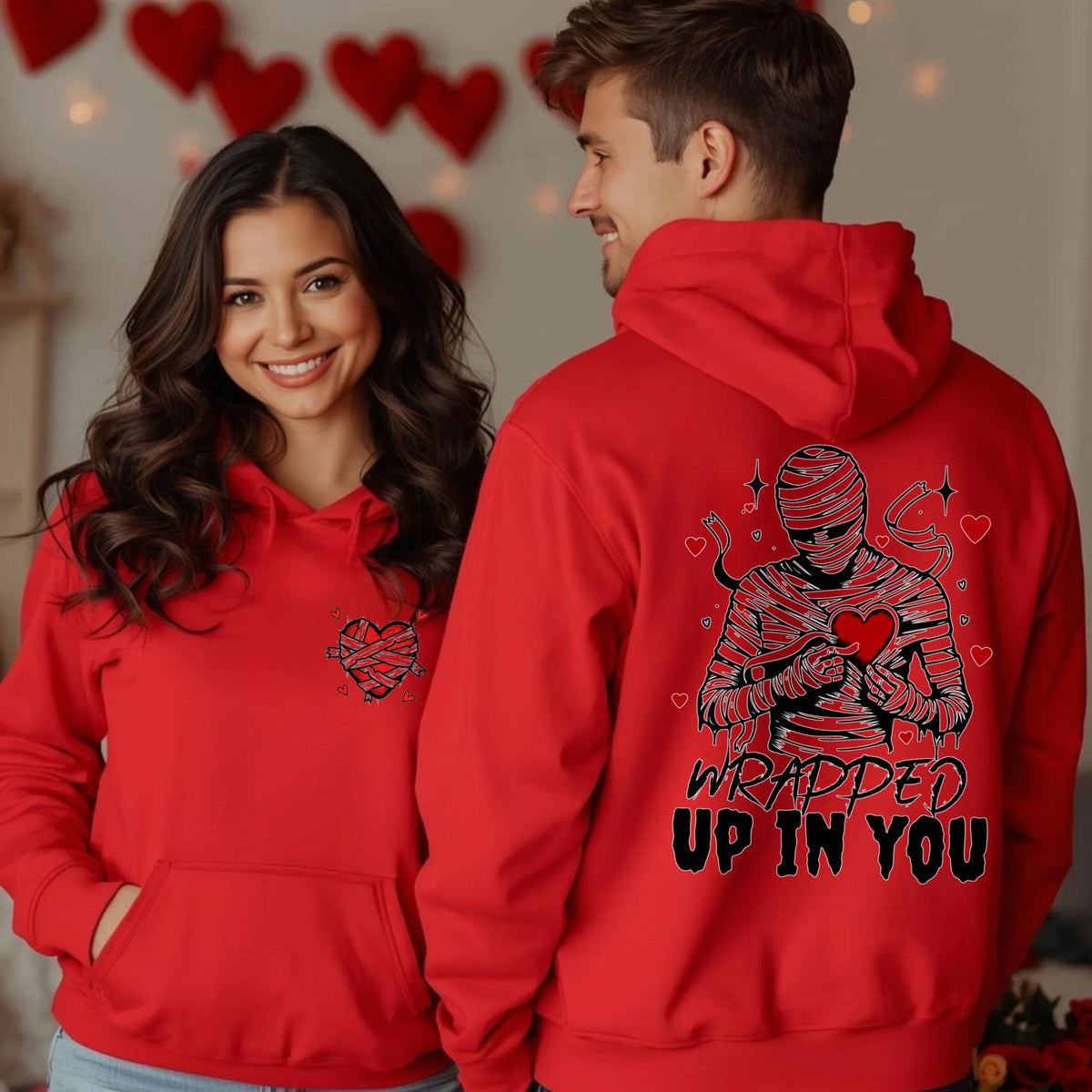 A smiling couple wearing bright red hoodies, one showing the wrapped heart front print and the other showing the mummy back graphic Wrapped Up In You.