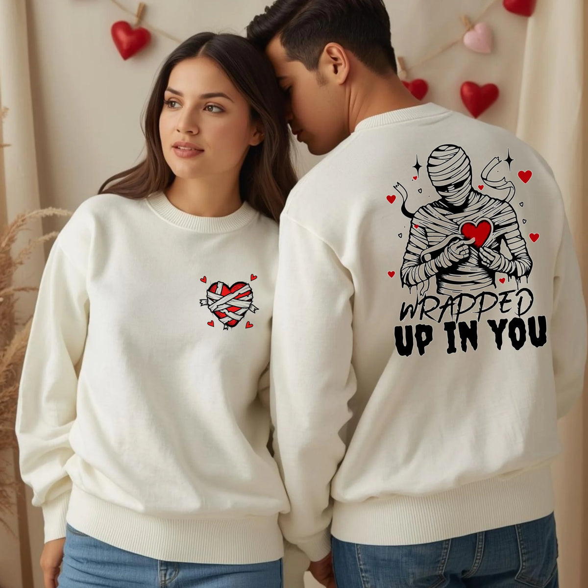 A cozy couple wearing white crewnecks featuring a wrapped heart front graphic and Wrapped Up In You mummy design on the back, cute Valentine matching sweater.