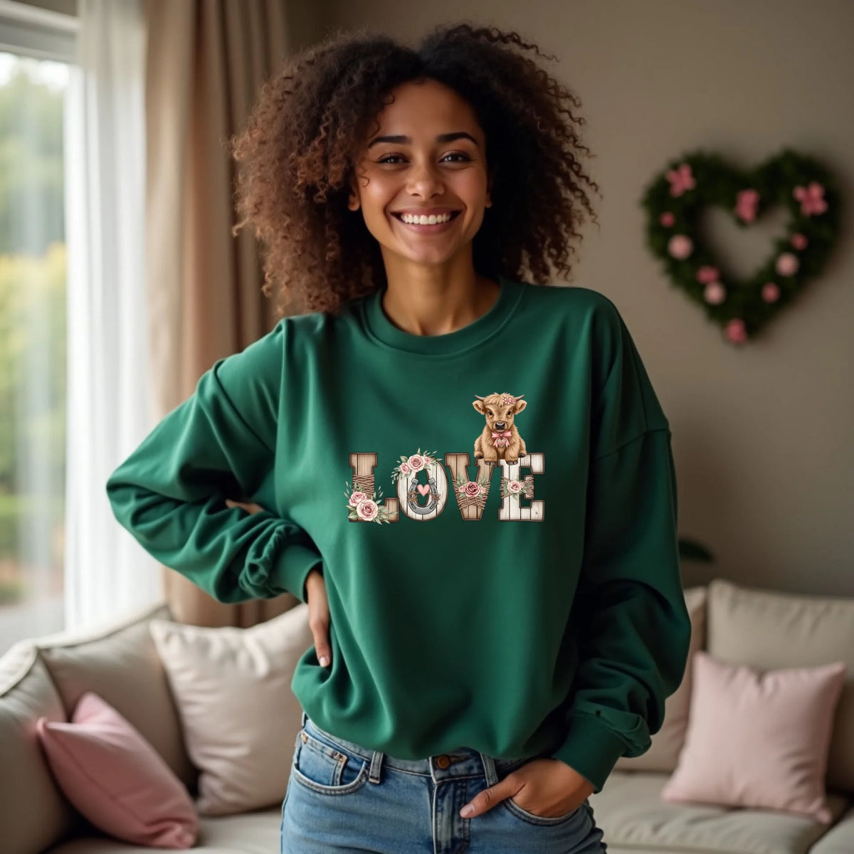 Model wearing a dark green crewneck with rustic wooden LOVE letters and floral calf art. Casual home setting with soft decor.