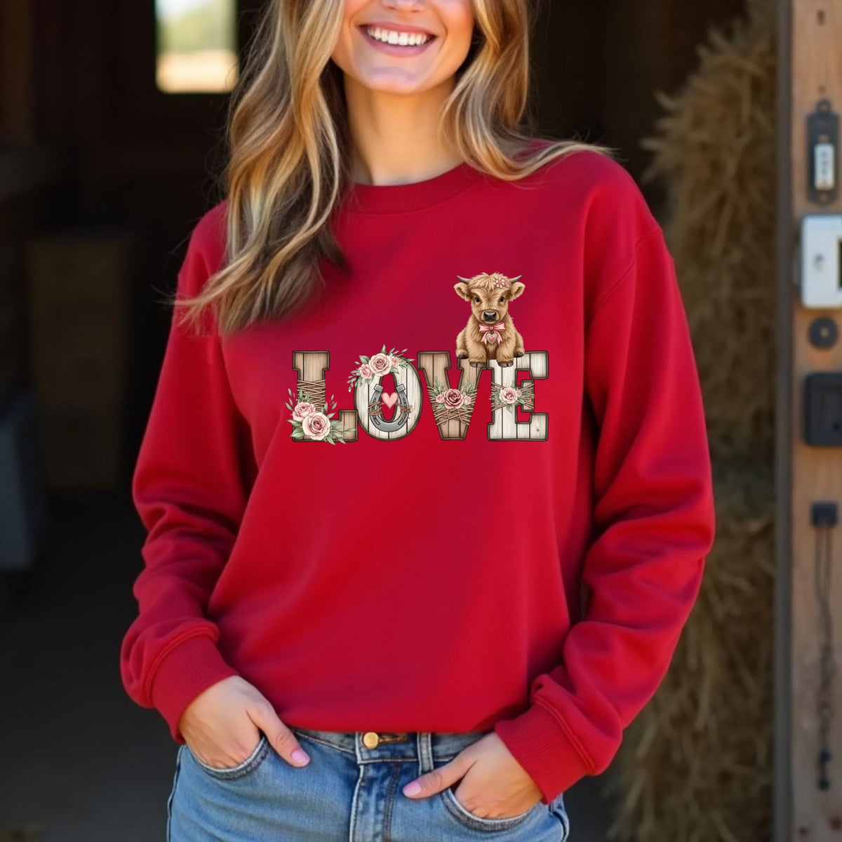 Woman wearing a red crewneck sweatshirt printed with a farmhouse-style LOVE design and highland calf artwork. Standing indoors in a warm rustic setting.