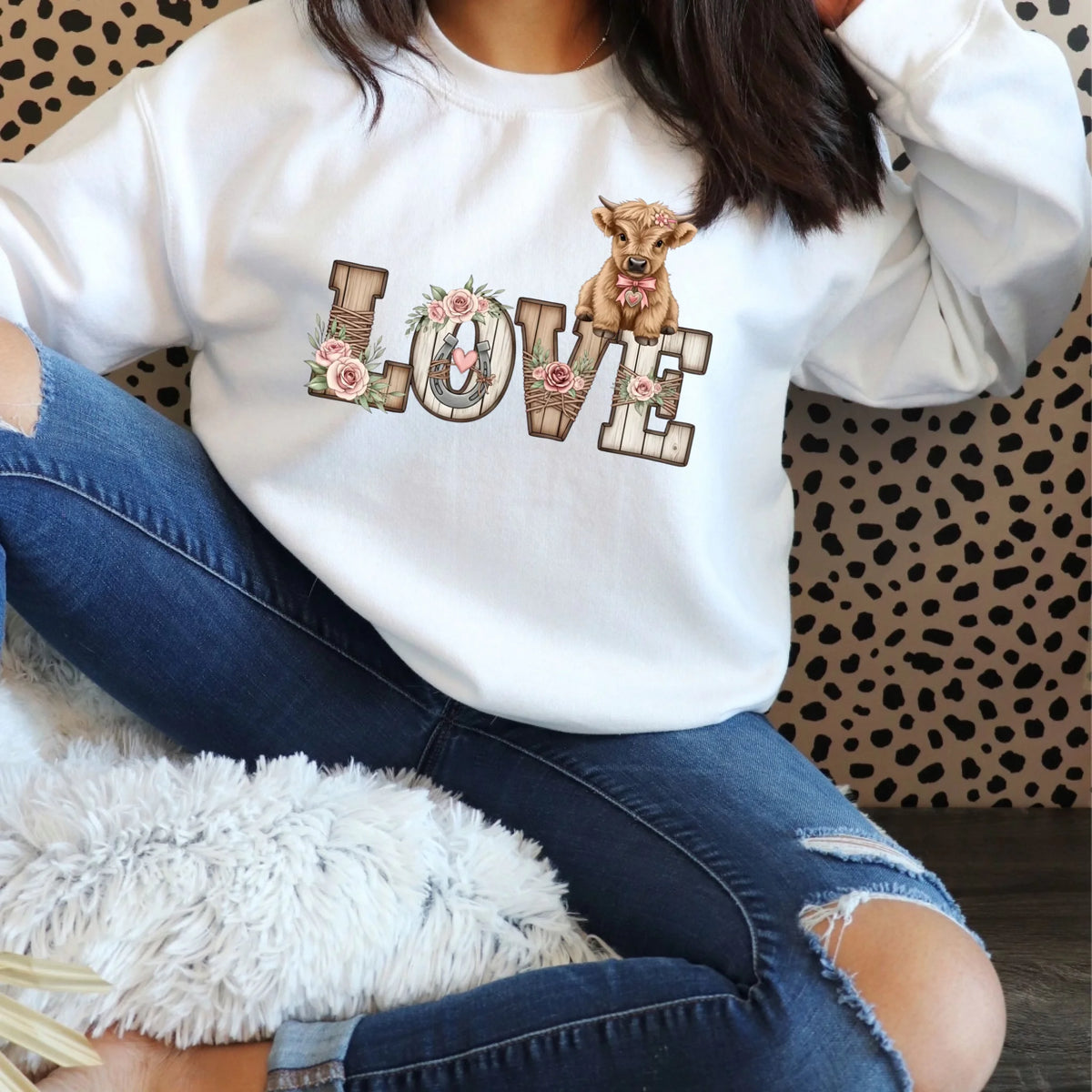 Woman wearing a white crewneck sweatshirt featuring a rustic wooden LOVE design with floral accents and a baby highland cow. Cozy indoor setting with casual seating.