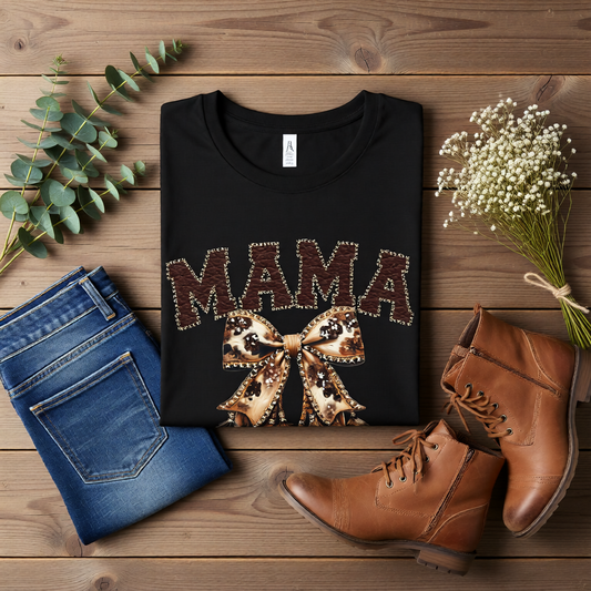 Flat lay of a black Bella Canvas tshirt mama cowhide bow shirt styled with denim jeans and boots on a rustic wooden background, featuring faux leather stitched mama lettering and a western bow graphic.