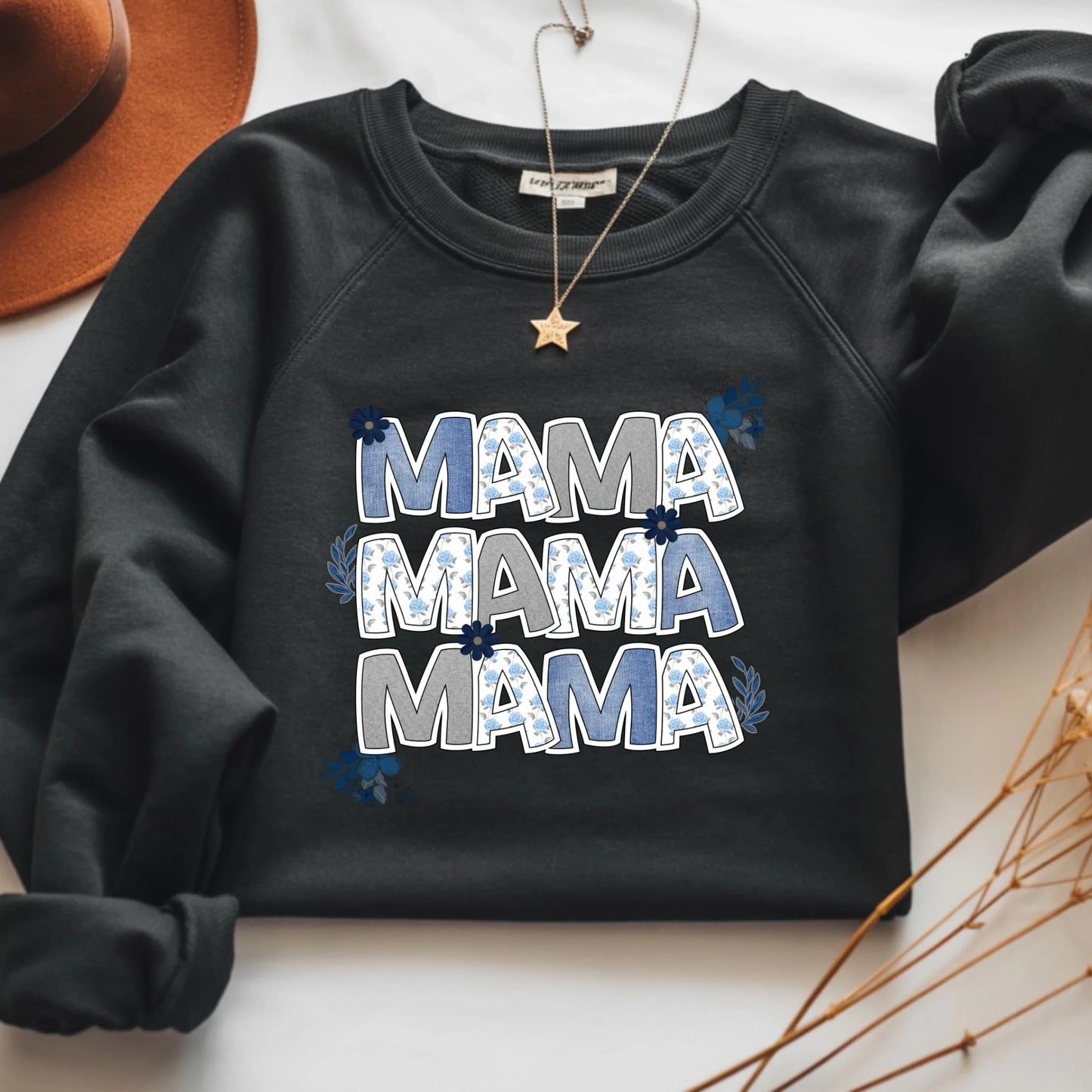 Flat lay of a black crewneck sweatshirt featuring a floral “Mama” letter design on a neutral background.