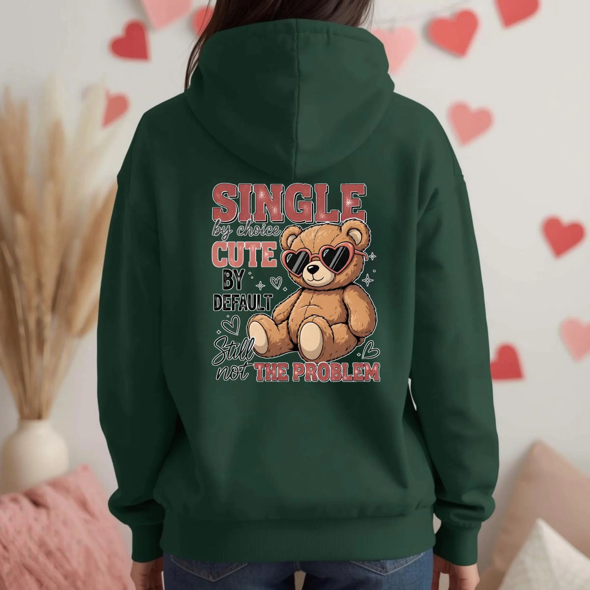 Person wearing a green hoodie with a teddy bear graphic and text, standing against a heart-patterned wall.