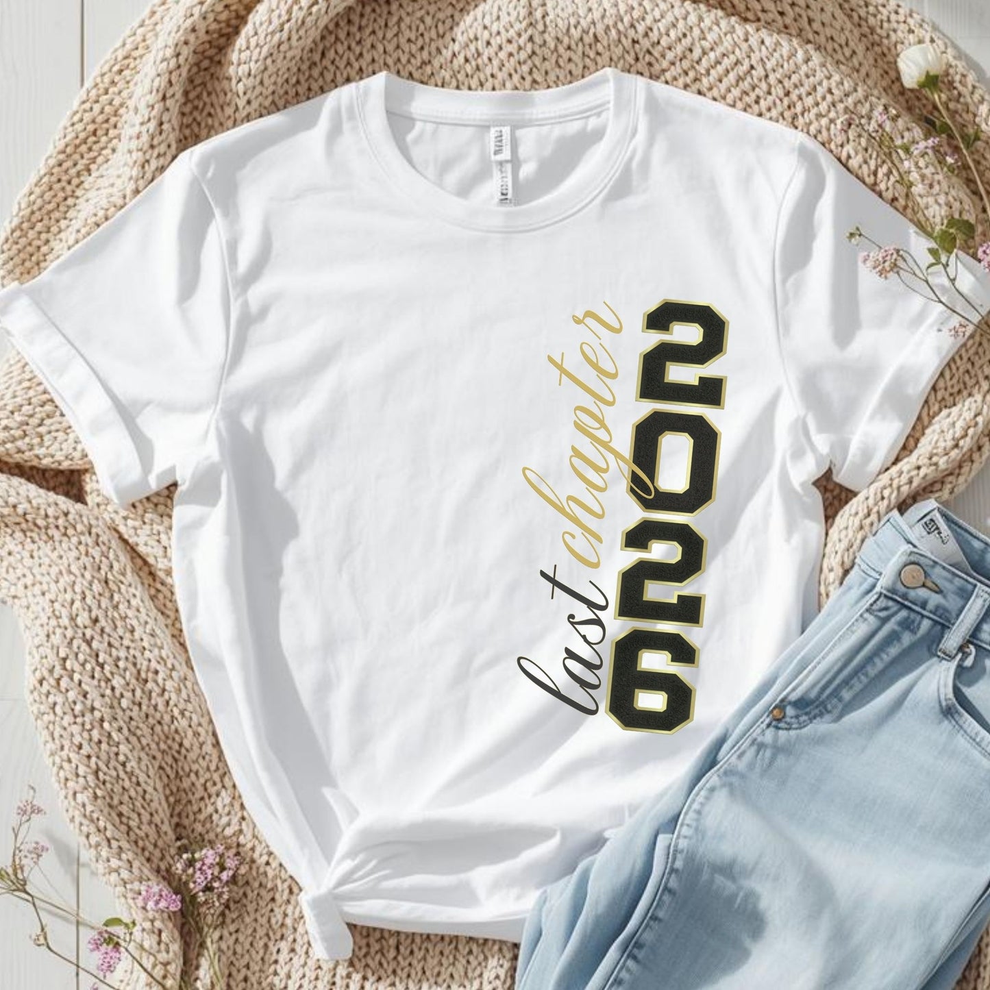 White t-shirt with '2026 last chapter' text on a textured surface with jeans and flowers.