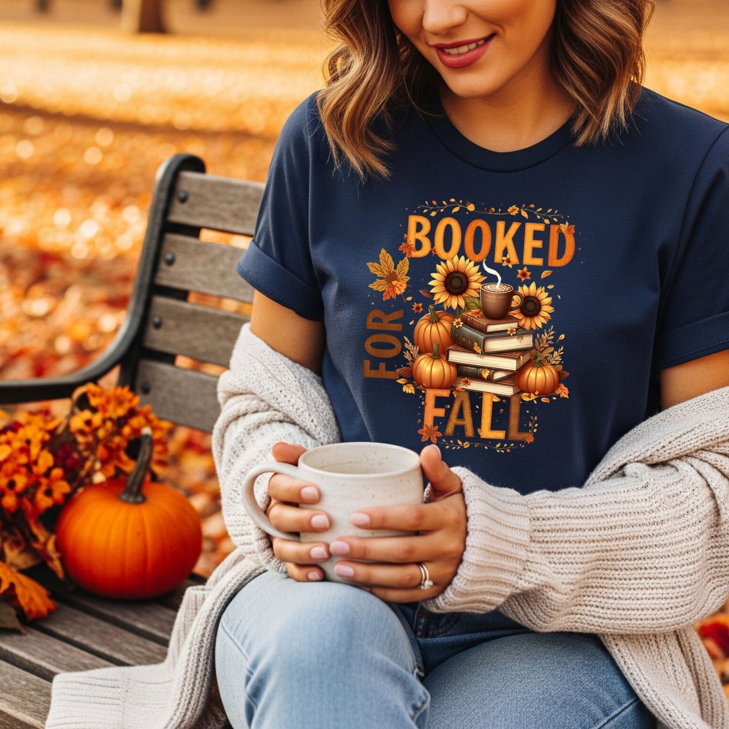Person wearing a 'Booked for Fall' shirt, holding a mug, sitting on a wooden bench with autumn decorations.
