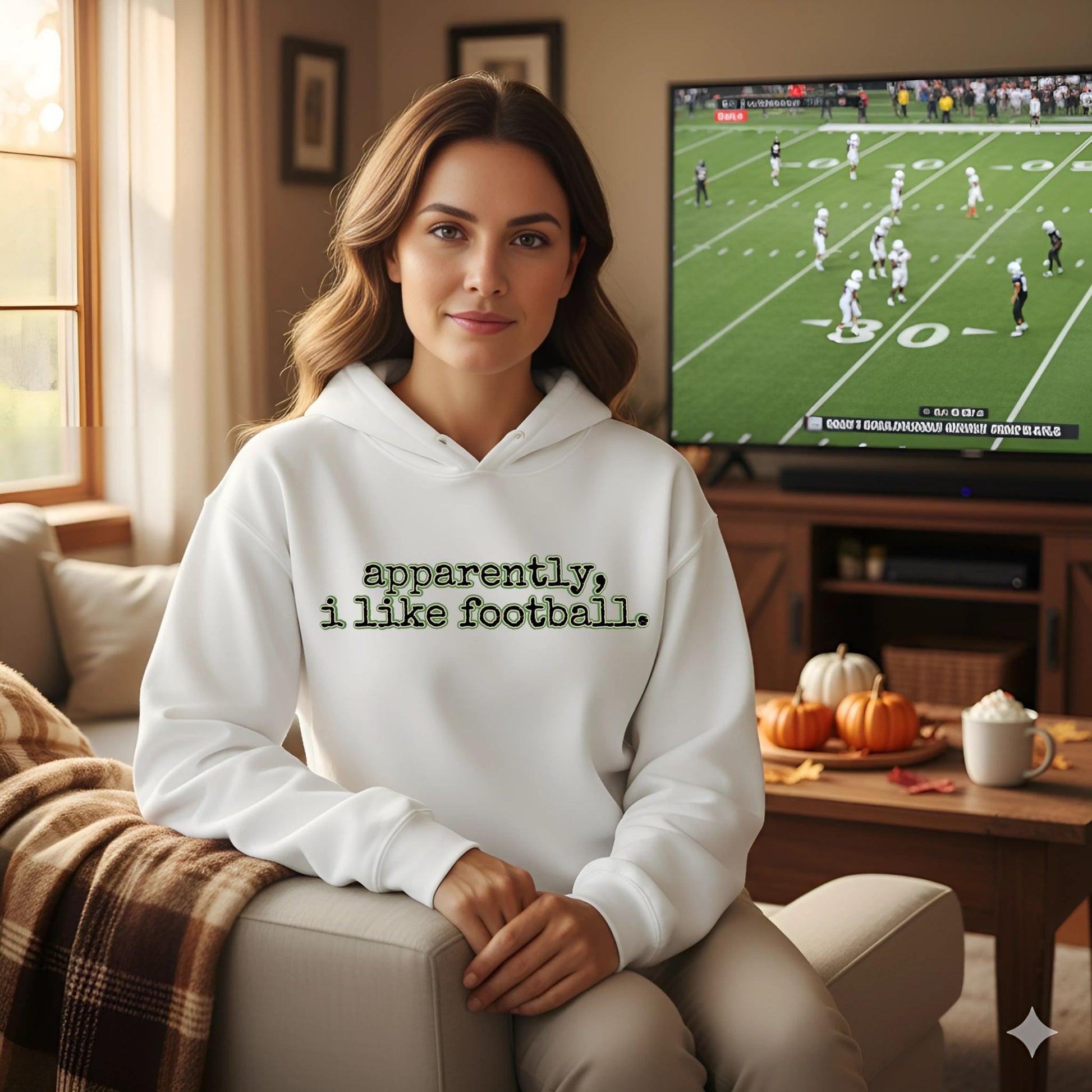 Woman wearing a hoodie with 'apparently, i like football.' text, sitting in a living room with a TV showing a football game.