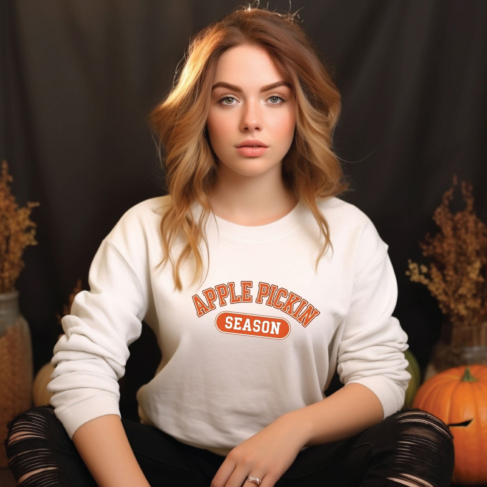 Woman wearing a 'Apple Pickin' Season' sweatshirt with pumpkins and fall decorations in the background.