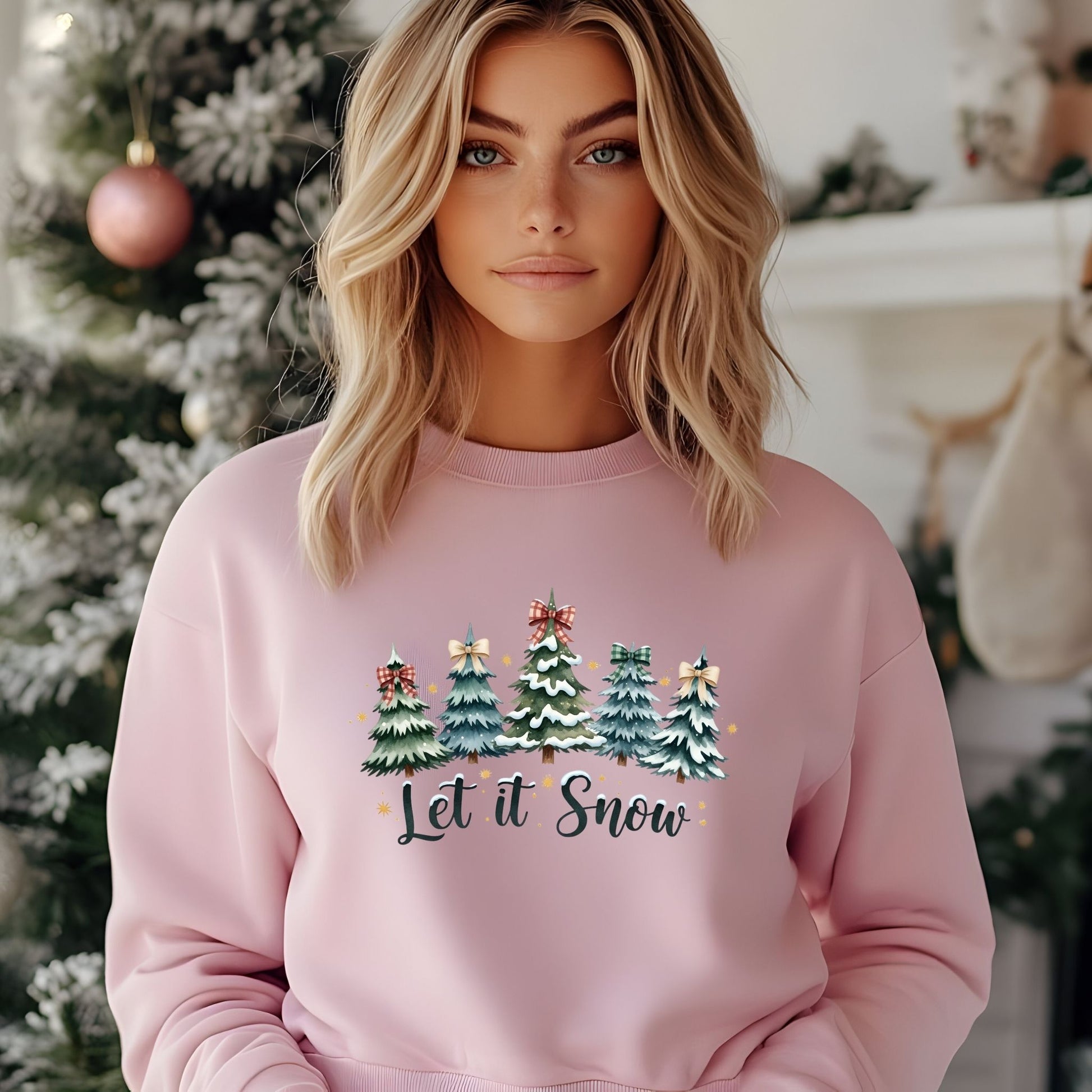 Woman wearing a pink sweatshirt with Christmas tree design and 'Let it Snow' text, standing in front of a decorated Christmas tree.