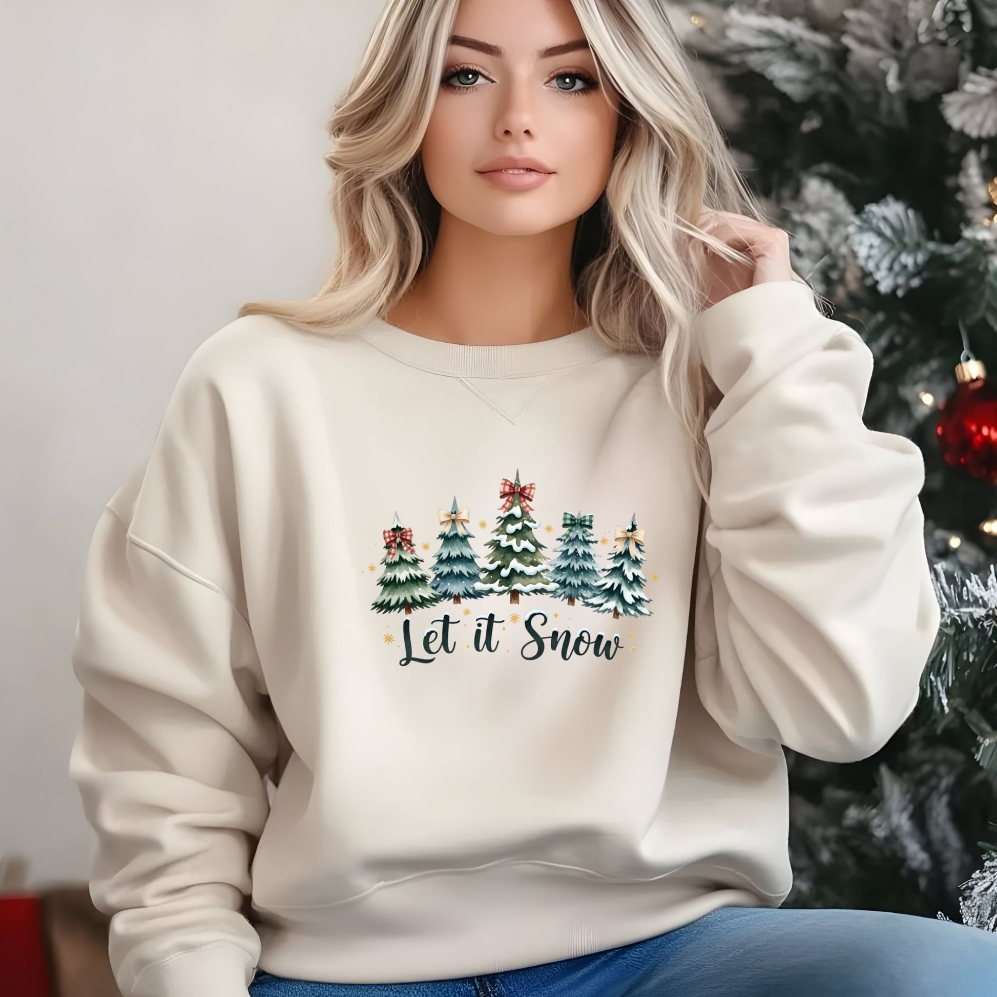 Woman wearing a cream sweatshirt with Christmas tree design and 'Let it Snow' text, standing in front of a decorated Christmas tree.