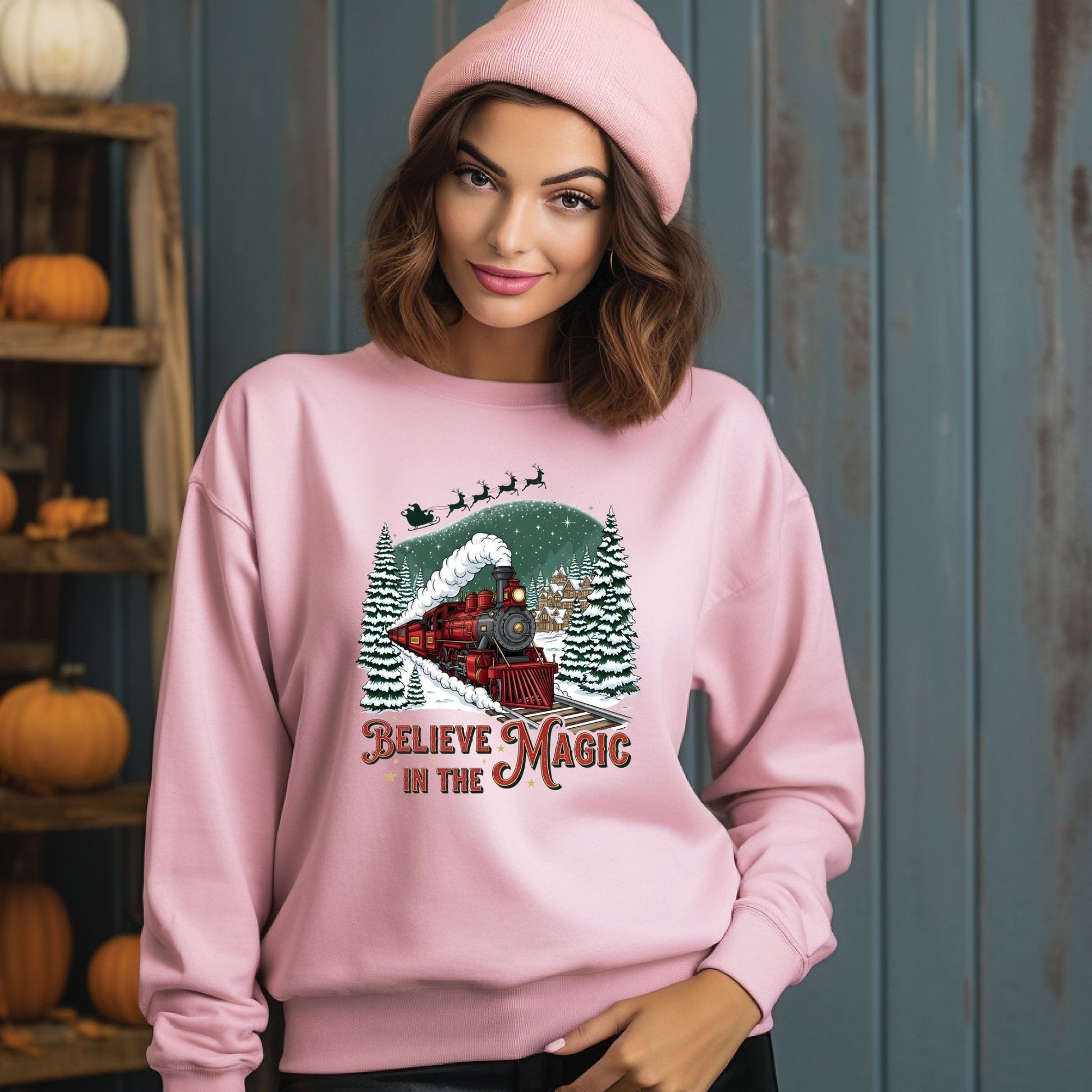Woman wearing a pink sweatshirt with a Christmas design and text, standing against a blue wall with pumpkins on shelves.
