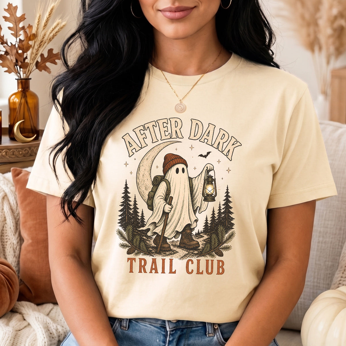 A woman wearing a cream-colored graphic t-shirt with a Halloween-themed design that reads 'AFTER DARK TRAIL CLUB' featuring a ghost in a sleeping hat and backpack holding a lantern in a forest.