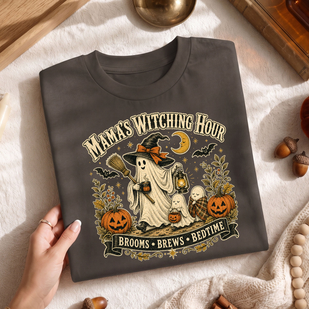 A folded dark charcoal gray graphic T-shirt is held by a hand over a cozy autumn flat-lay background. The shirt features a detailed Halloween-themed graphic with a ghostly witch, a small ghost, jack-o'-lanterns, bats, a crescent moon, and the text 'MAMA'S WITCHING HOUR' and 'BROOMS • BREWS • BEDTIME'.