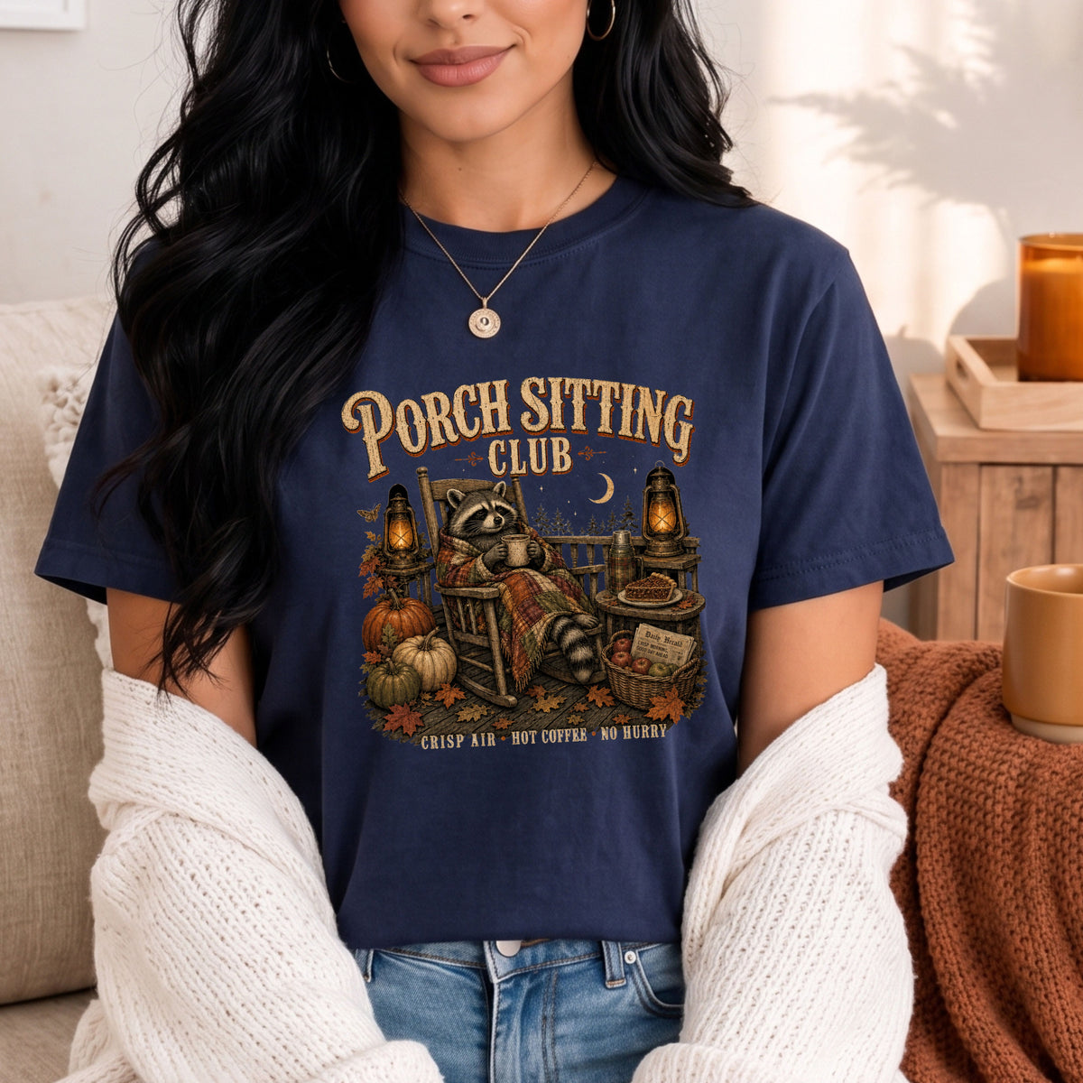 A woman wearing a navy blue graphic t-shirt with a 'Porch Sitting Club' fall-themed raccoon illustration, styled with a white knit cardigan and blue jeans in a cozy indoor setting.