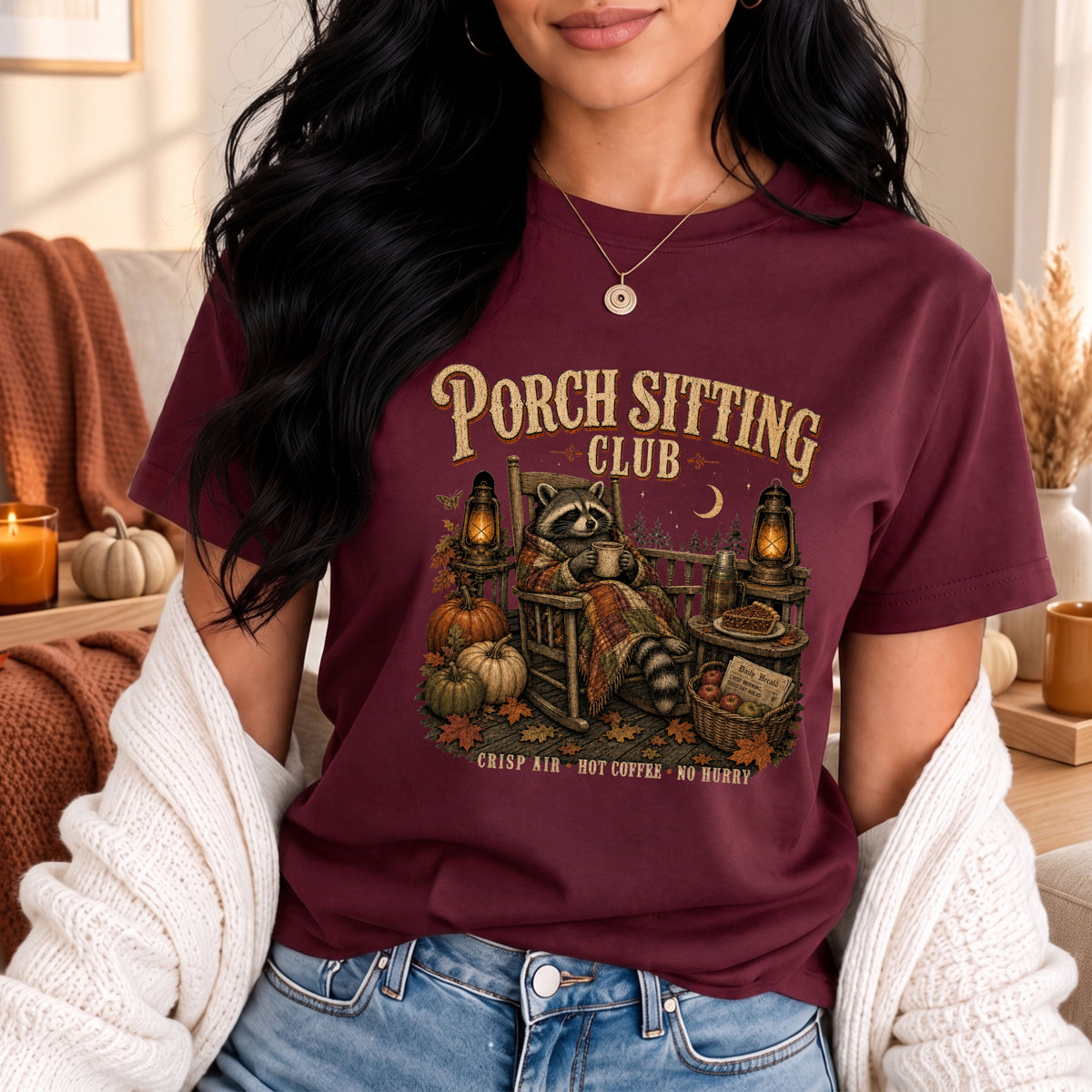 A woman wearing a burgundy graphic t-shirt with a 'Porch Sitting Club' autumn-themed raccoon illustration, styled with a white knit cardigan and blue jeans in a cozy, warm-toned home setting.
