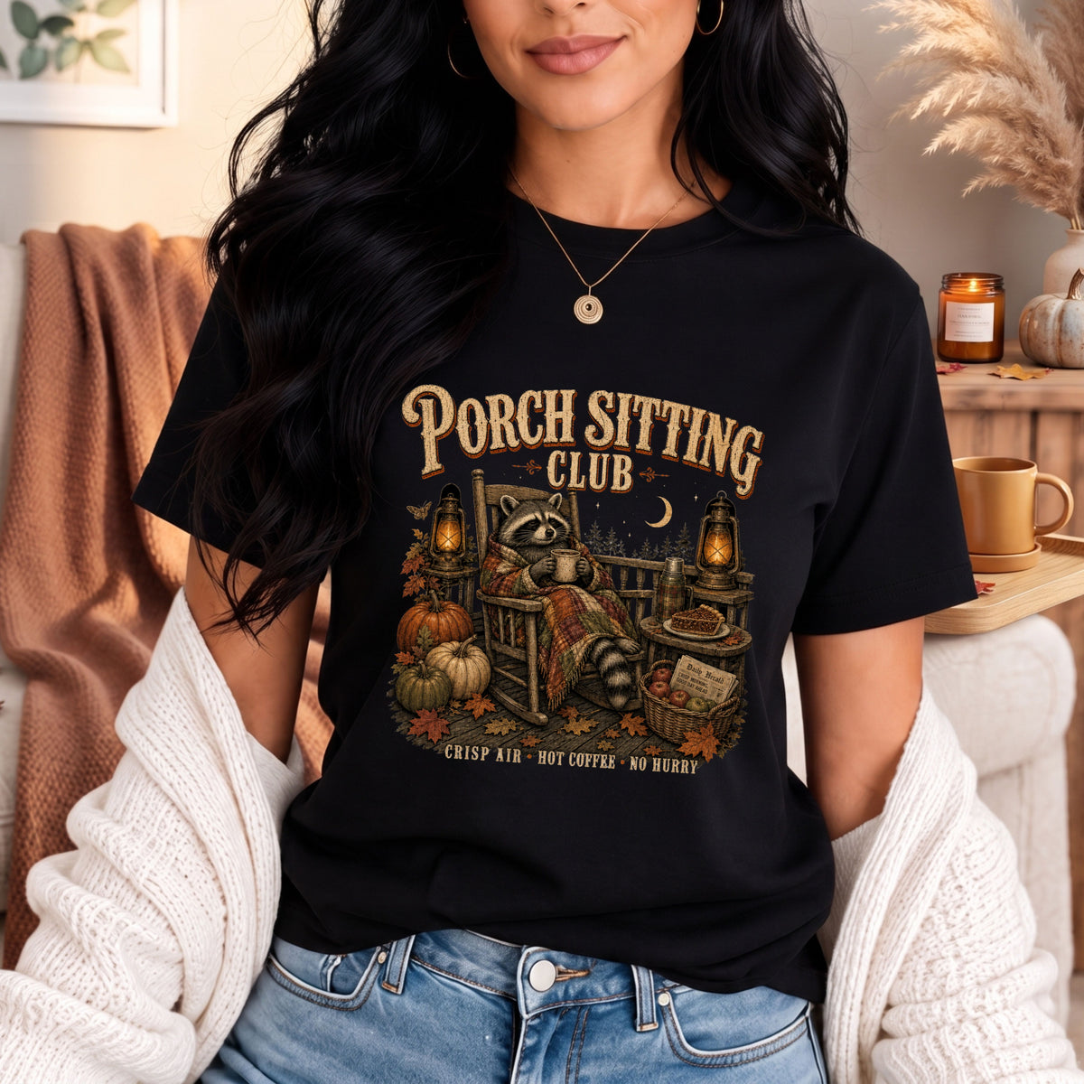 A woman wearing a black graphic t-shirt with a fall-themed 'Porch Sitting Club' design, styled with a white knit cardigan and blue jeans in a cozy indoor setting.
