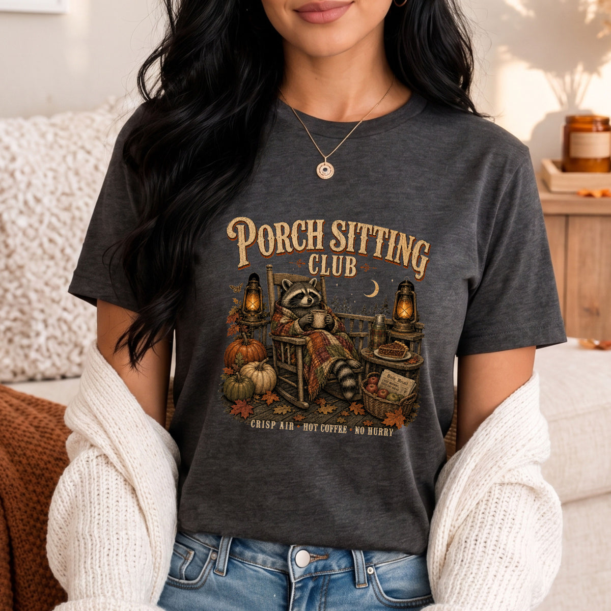 A woman wearing a dark charcoal heather graphic t-shirt with a 'Porch Sitting Club' fall-themed illustration, styled with a white knit cardigan and blue jeans in a cozy indoor setting.