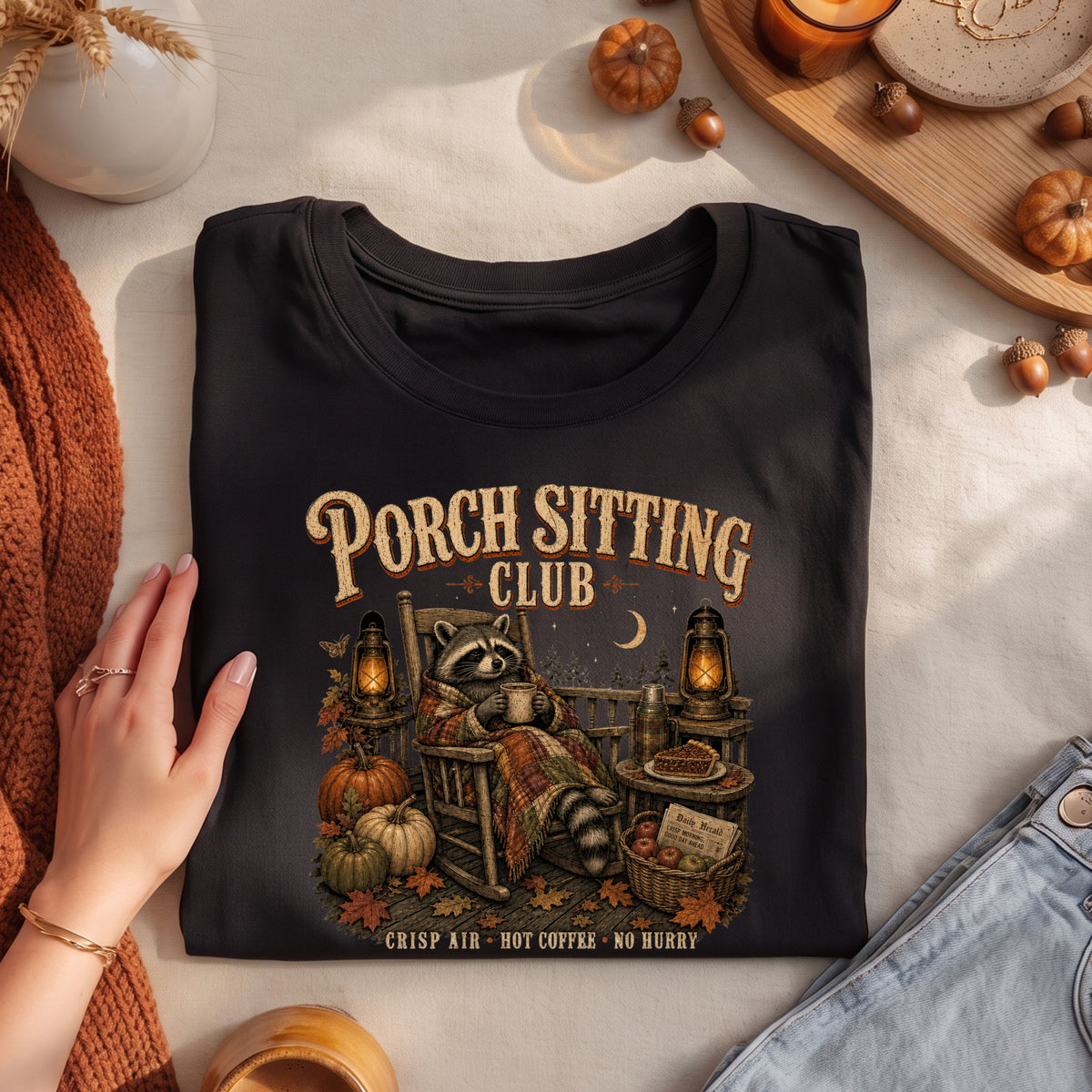 A folded black graphic T-shirt is styled in a flat lay with autumn props. The shirt features a raccoon sitting in a porch chair by a window with pumpkins, lanterns, and the text 'PORCH SITTING CLUB' and 'CRISP AIR - HOT COFFEE - NO HURRY'.