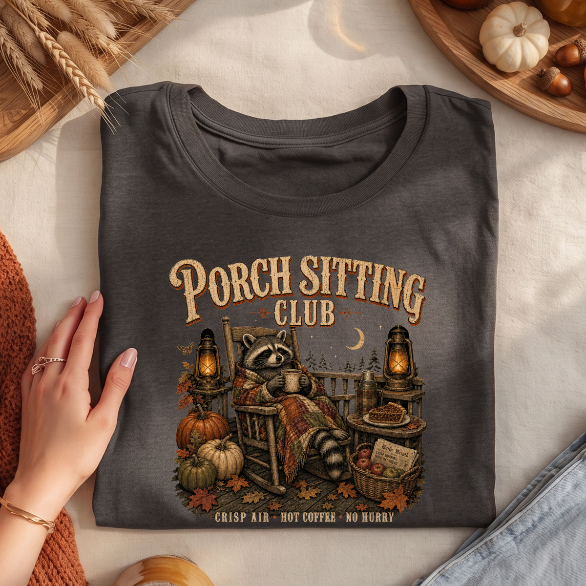 A folded dark charcoal gray graphic T-shirt is displayed on a light fabric surface with seasonal props. The shirt features a printed illustration of a raccoon sitting in a rocking chair by a porch, surrounded by pumpkins, lanterns, and autumn decor, with the text 'Porch Sitting Club' and 'CRISP AIR • HOT COFFEE • NO MERRY'.