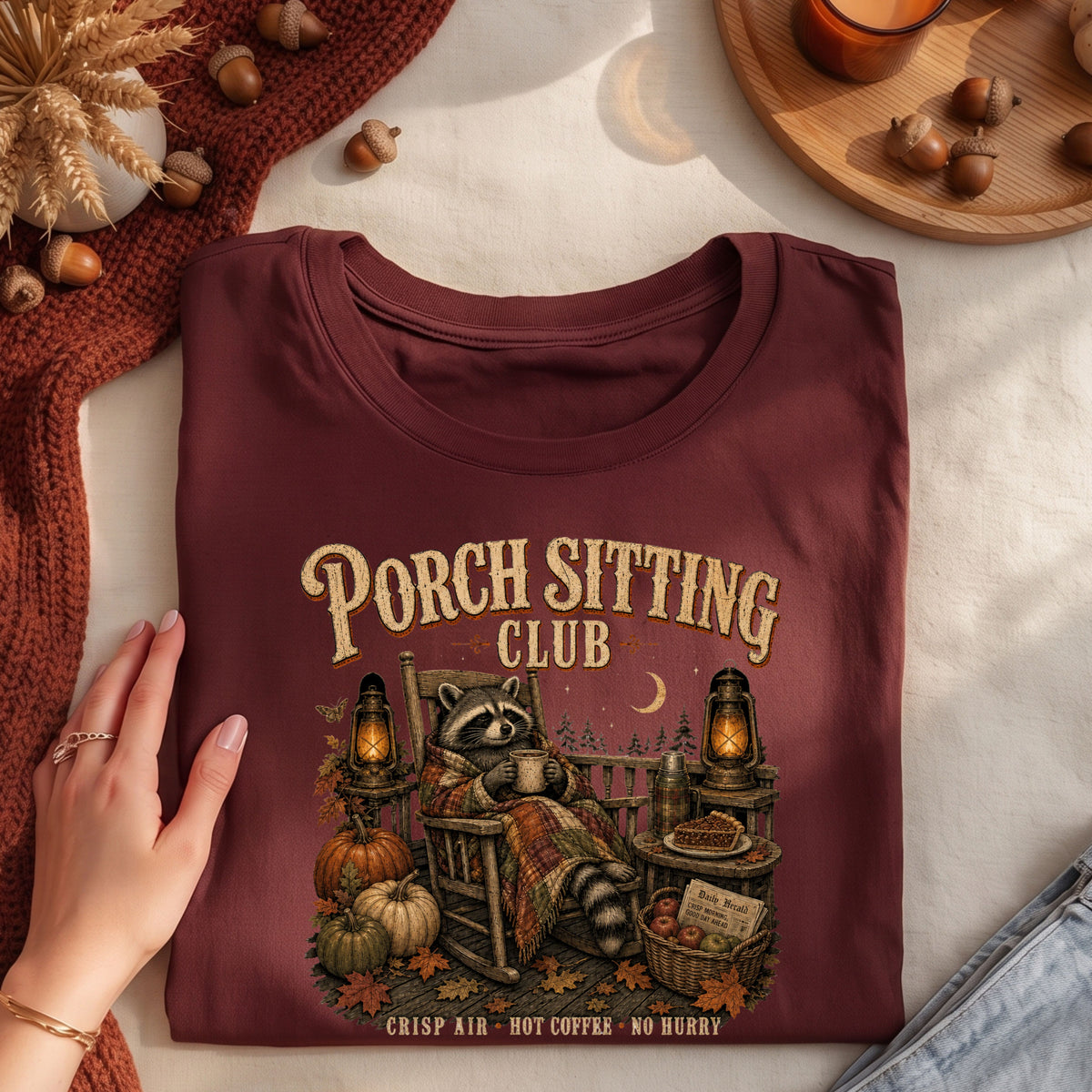 A folded maroon graphic T-shirt is styled on a light fabric surface with autumn props. A hand rests on the left side of the shirt.