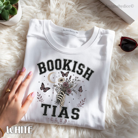 A folded white graphic T-shirt laid on a fluffy cream faux-fur surface, with a hand resting on the left side. The shirt has a large front print reading “BOOKISH TIAS CLUB” with butterflies, a crescent moon, and floral artwork.