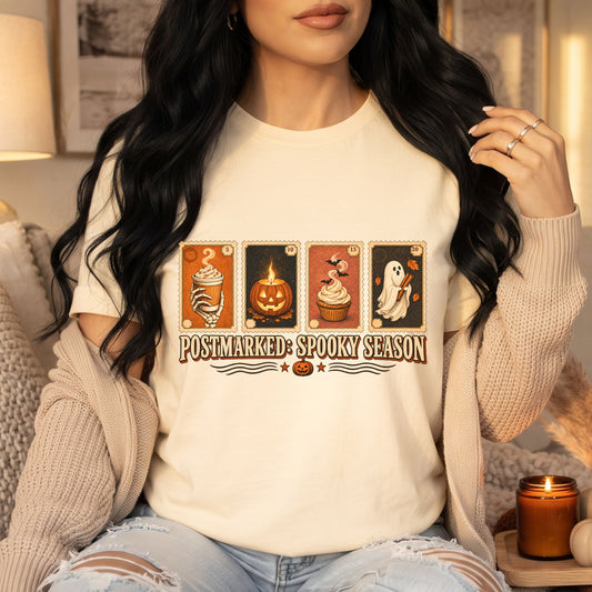 A woman wearing a cream-colored graphic t-shirt with a 'Postmarked Spooky Season' Halloween card design, styled with a beige knit cardigan in a cozy indoor setting.