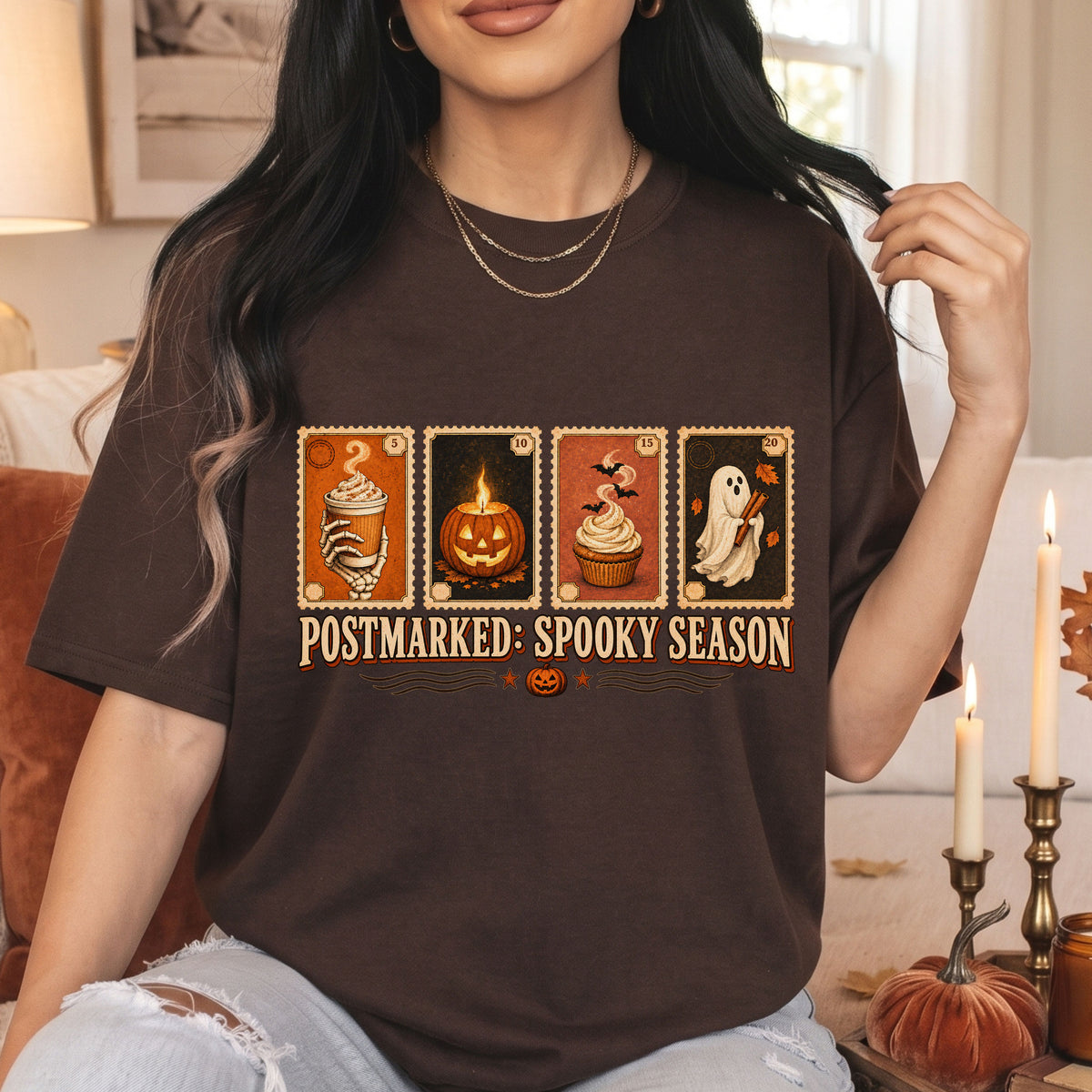 A woman wearing a dark brown graphic t-shirt with a Halloween-themed stamp design and the text 'POSTMARKED: SPOOKY SEASON'. The background shows a cozy indoor setting with candles, a pumpkin, and a sofa.