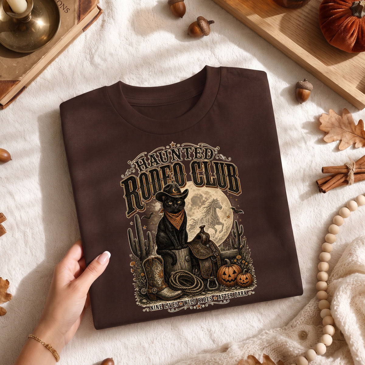 A folded dark brown graphic T-shirt is held by a hand over a cozy autumn-themed fabric surface. The shirt features a detailed western-themed print of a cat in a cowboy hat with the text 'Haunted Rodeo Club' and a moonlit scene with cacti, boots, and pumpkins.
