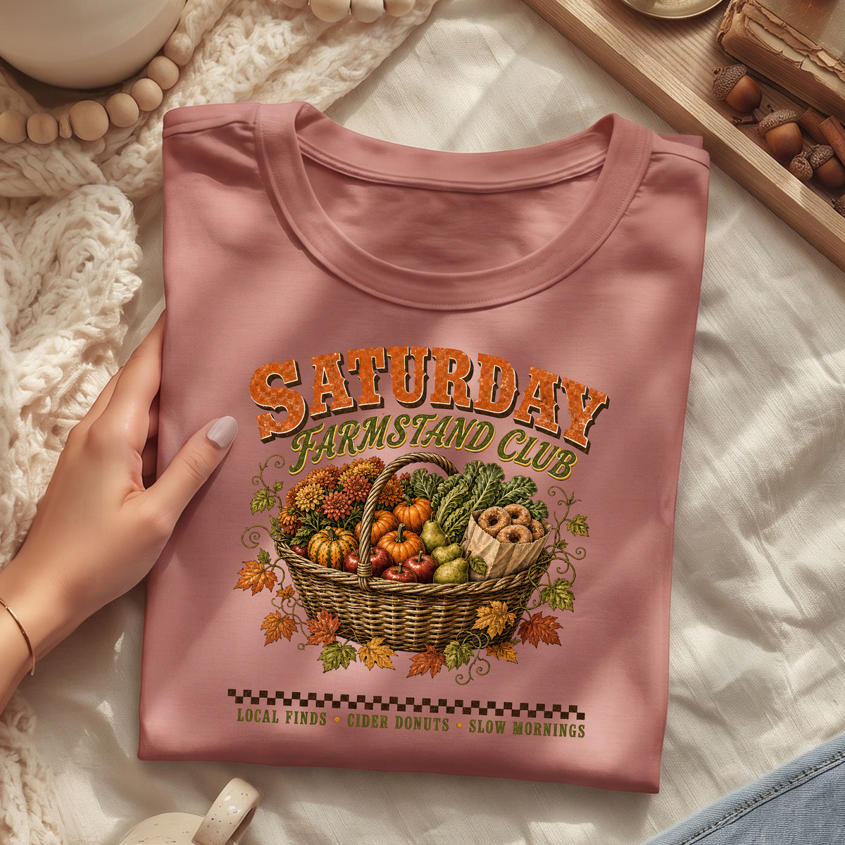A folded dusty rose t-shirt with a 'Saturday Farmstand Club' graphic print, held by a hand over a cozy white blanket background with autumn-themed props.