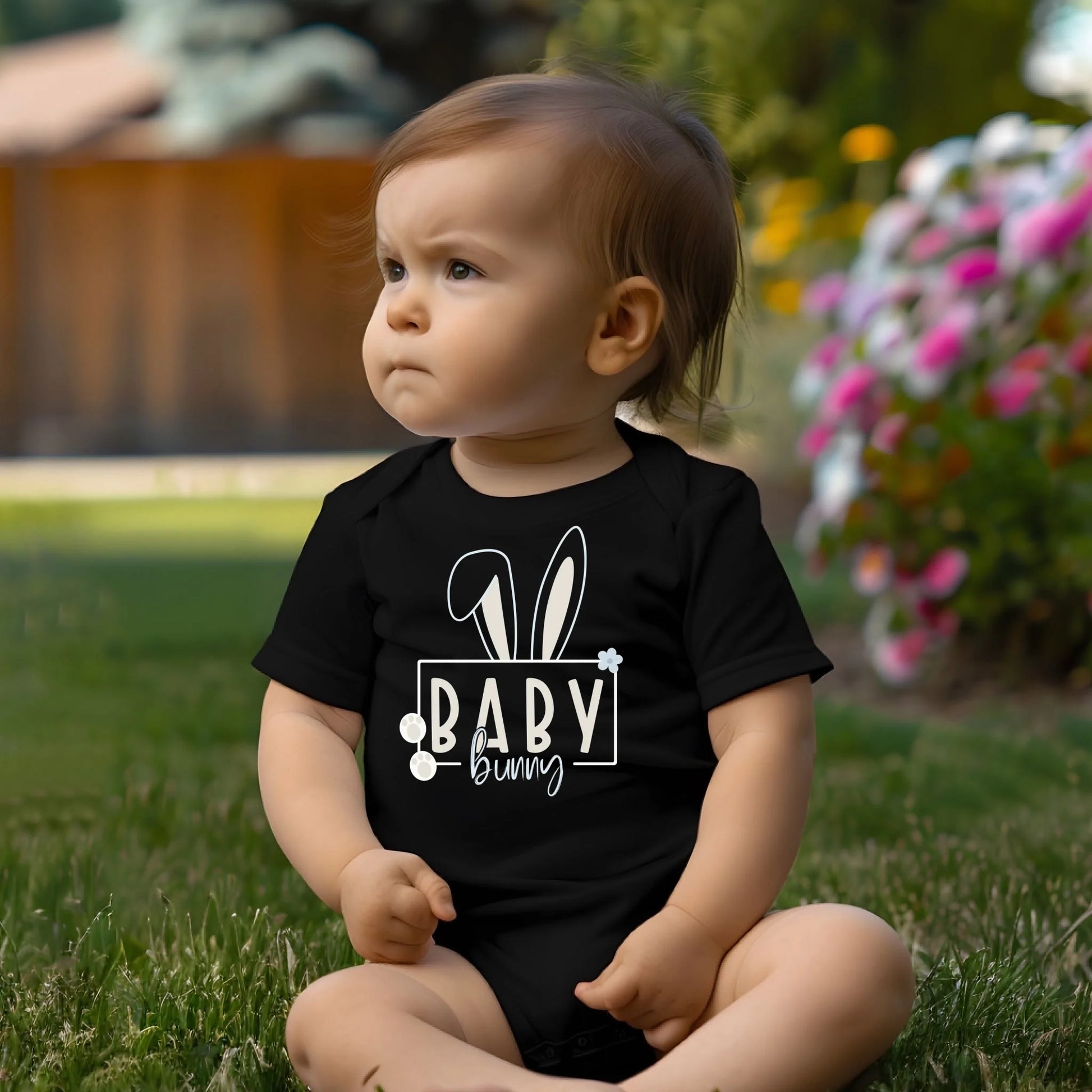 Infant wearing a black Easter bunny baby onesie sitting outdoors in a natural spring setting.