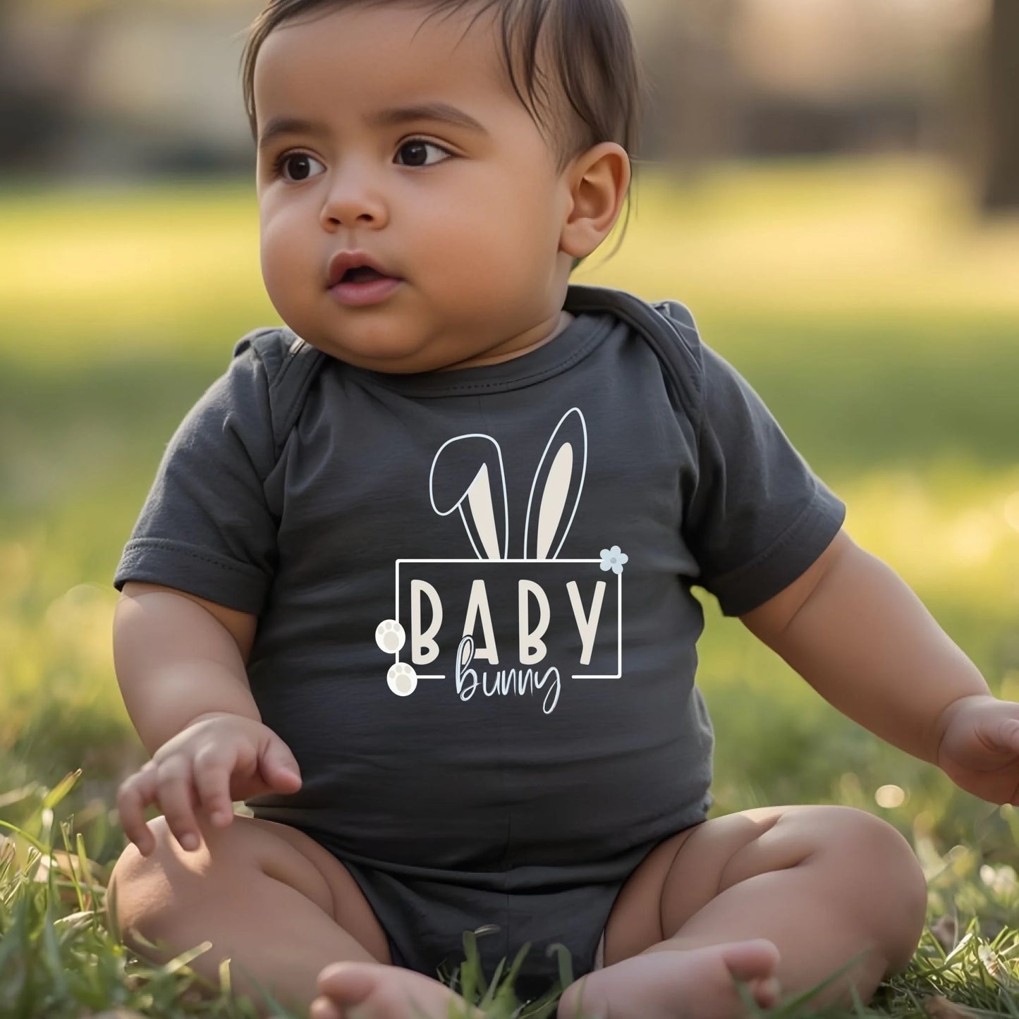 Infant wearing a gray Easter bunny baby onesie while sitting outdoors in natural daylight.