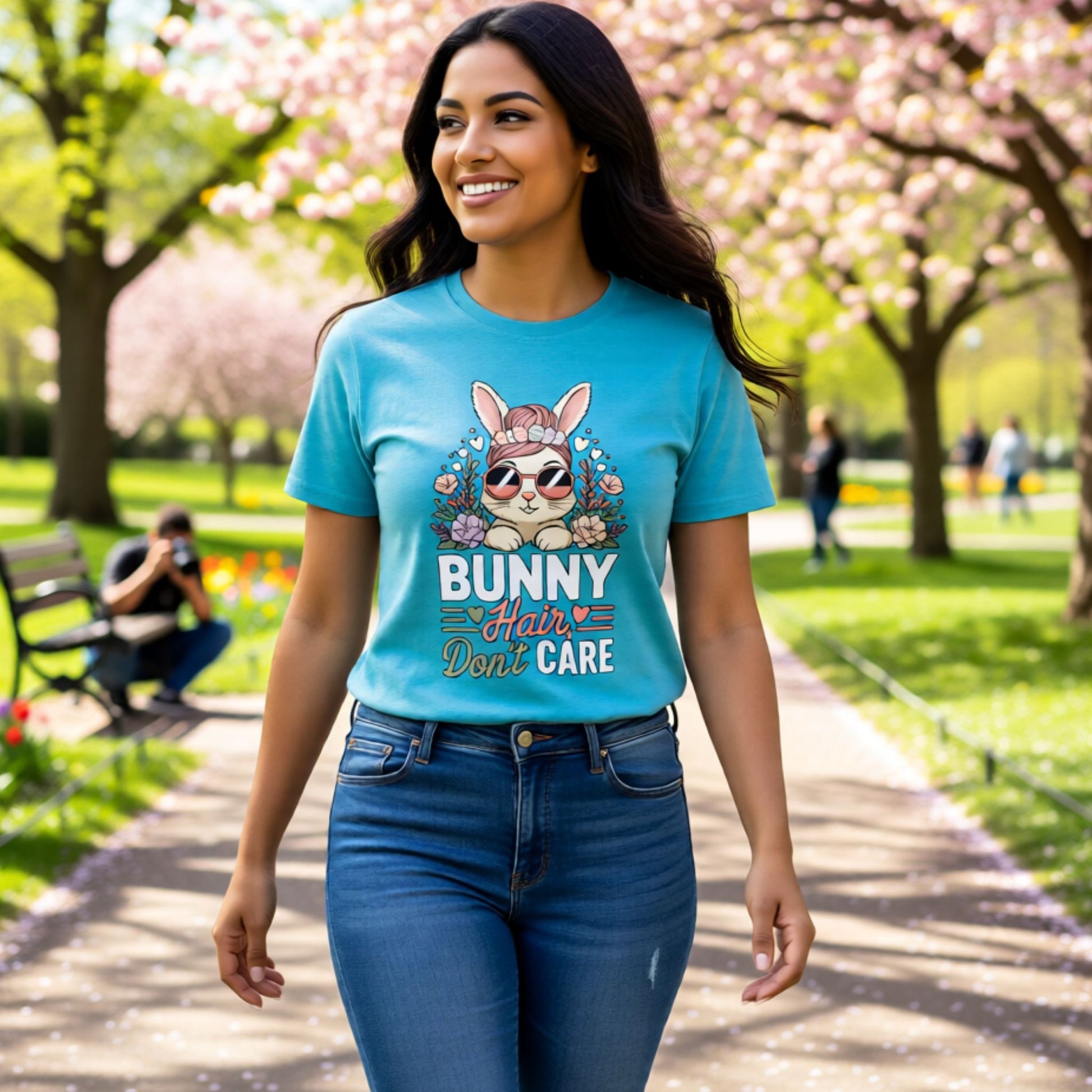 Woman wearing a aqua t-shirt with a bunny graphic and text in a park with cherry blossoms.