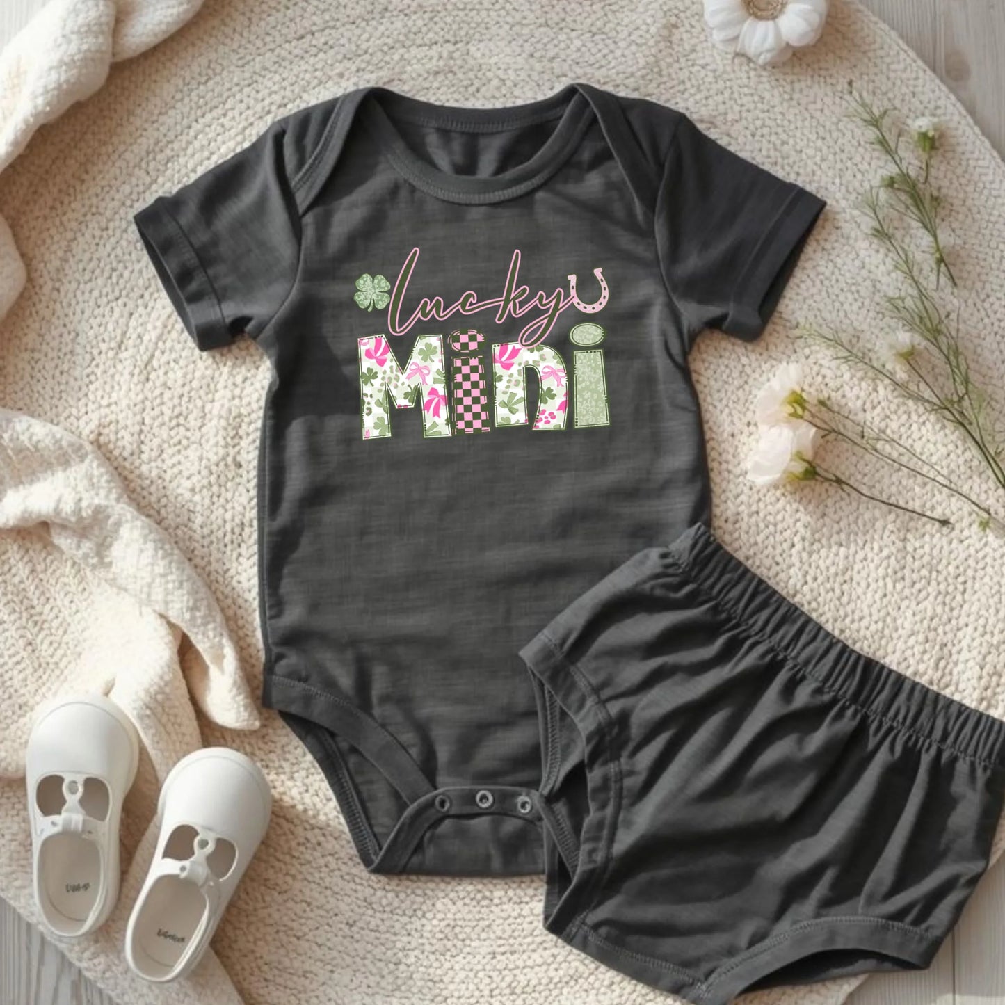 dark grey colored  baby onesie with 'Lucky Mini' text and matching shorts on a soft surface with white shoes and flowers.